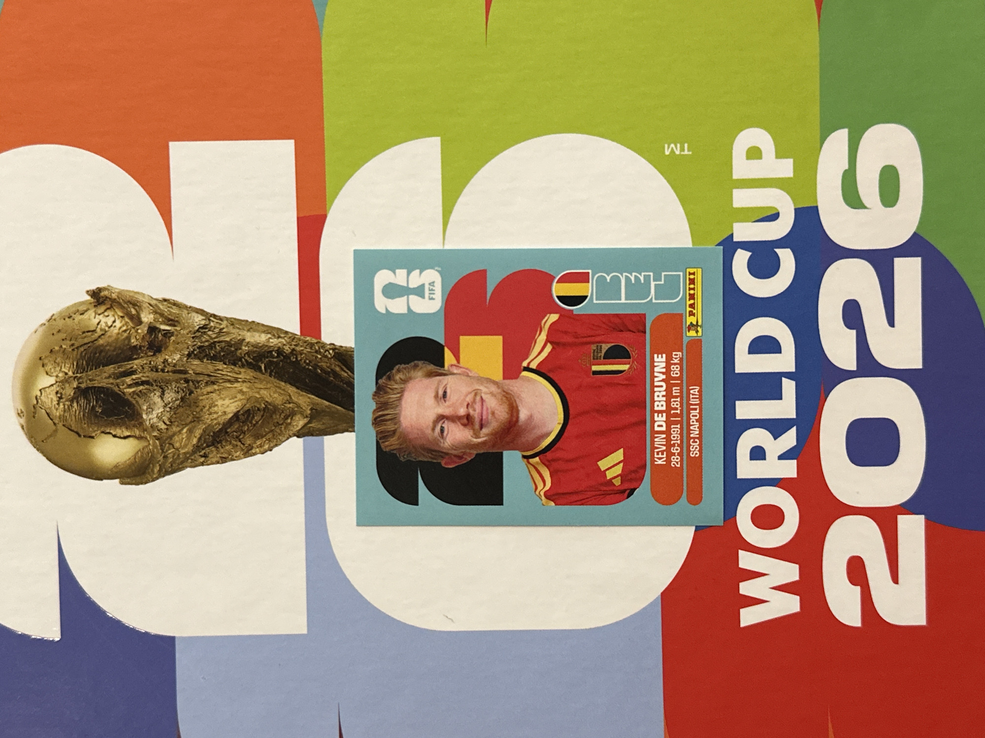 The image displays a detailed view of a colorful poster or page, prominently featuring a FIFA World Cup 2026 theme. At the top, a golden replica of the FIFA World Cup trophy stands, positioned centrally and partially overlapping a large white '2' numeral in the background. Directly below the trophy, a rectangular Panini sticker of Belgian footballer Kevin De Bruyne is affixed to the page.
The sticker, with rounded corners, is primarily light blue and depicts De Bruyne smiling while wearing a red Belgium national team jersey, which includes a yellow Adidas logo on the left chest and a black, yellow, and red stripe representing the Belgian flag on the right sleeve. Below his portrait, white text on two orange horizontal bars provides his name "KEVIN DE BRUYNE", birthdate, height, and weight ("28-6-1991 | 1,81 m | 68 kg"), and club information "SSC NAPOLI (ITA)". A small Panini logo is visible in the bottom right corner of the sticker. The sticker also incorporates abstract design elements, including a stylized "23" in black at the top left, a FIFA logo above it, and vertical "FIFA WORLD CUP" text alongside a Belgian flag graphic on the right side.
The background of the image features large, abstract white numerals forming "2026" against blocks of color. The upper '2' is on a blue background, the '0' on an orange background, the lower '2' on a red background, and the '6' on a green background. The words "WORLD CUP" are printed in large, bold white capital letters above the lower "2026". A trademark symbol (TM) is subtly visible on the right side, within the green section of the background. The poster's color palette includes distinct blocks of blue, orange, red, and green. This scene is situated in the city of Prisovo, Bulgaria.