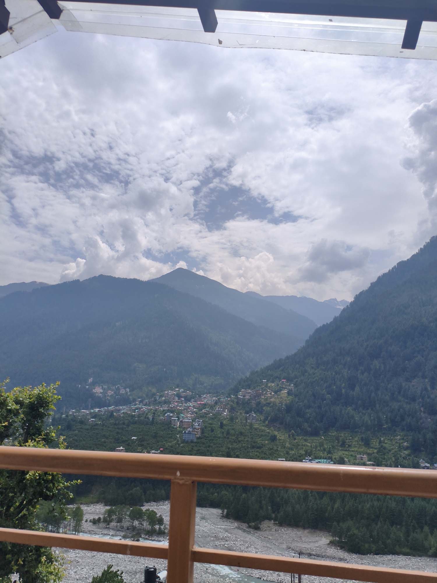 The image depicts a mountainous landscape with a village nestled in a valley. The foreground features a brown railing, and beyond that, a riverbed filled with rocks and a flowing river. Trees line the river and climb up the slopes of the mountains. A village with buildings of various colors is spread across the lower part of the mountainside. The sky is filled with scattered clouds. The scene is likely the city of Vashisht, India, given the architecture and mountainous terrain, which are characteristic of the region.