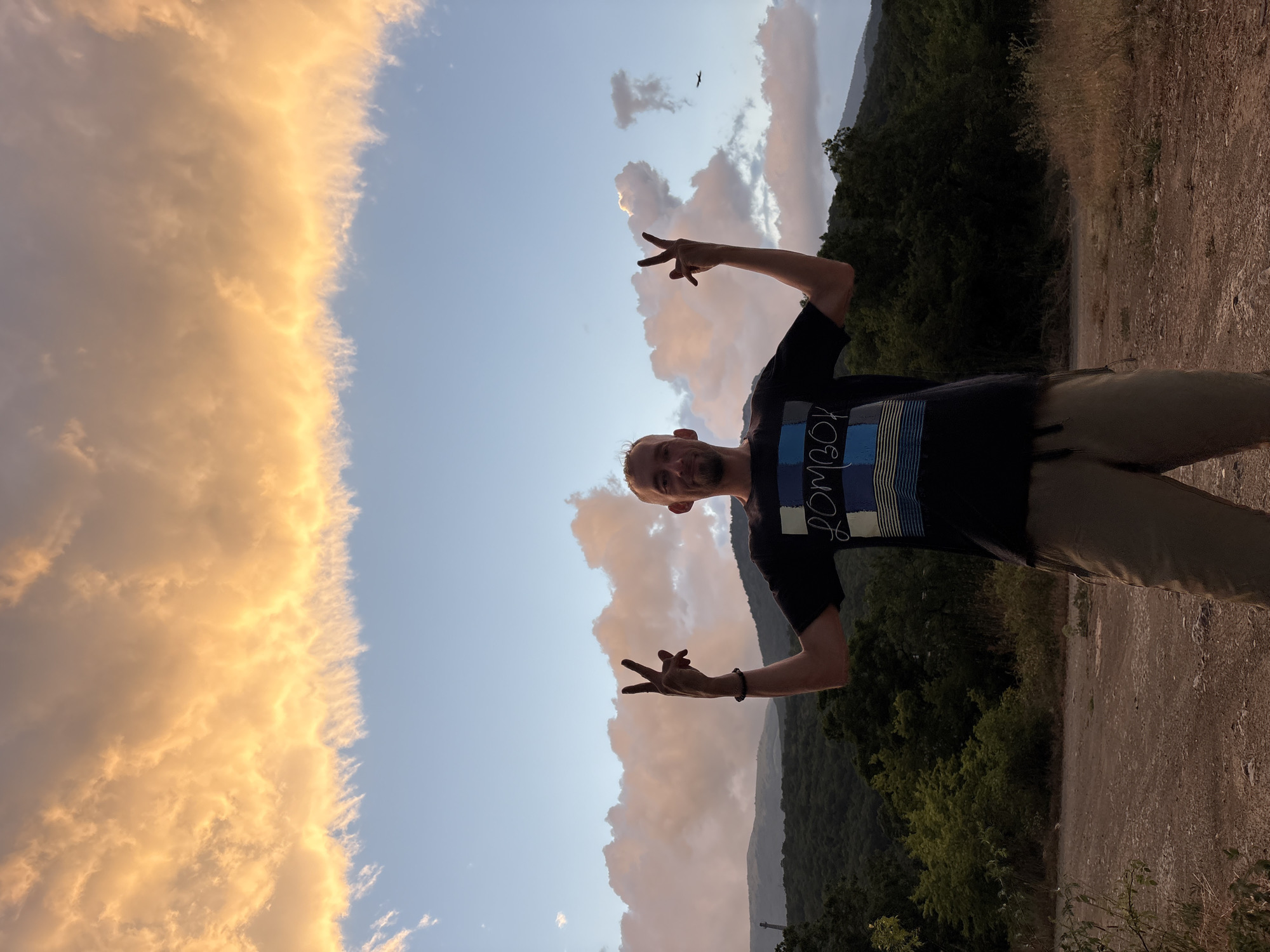 A man stands in a dirt lot, silhouetted against a dramatic sky filled with orange and pink clouds and patches of blue. He is wearing a dark t-shirt with a blue and white checkered pattern and the word "LOMBOK" printed in a stylized font, along with horizontal stripes. He is also wearing khaki shorts. He has his arms raised, and his hands are forming peace signs. Behind him, rolling green hills covered in trees stretch towards the horizon, with a hazy, mountainous landscape in the distance. The light suggests it is either sunrise or sunset. The specific location in Malyi Mayak, Ukraine is not identifiable from the visual information.