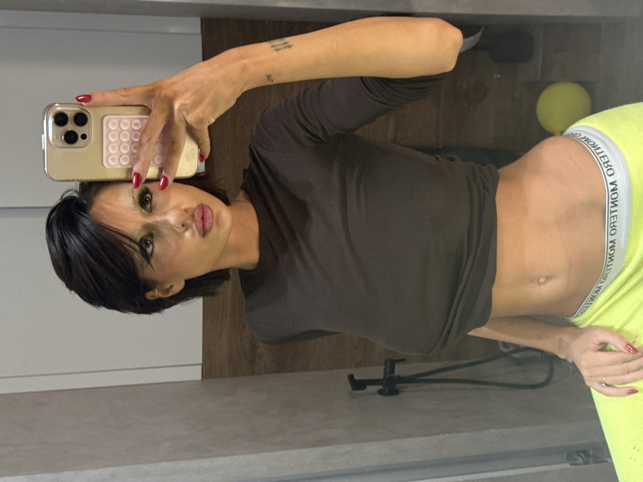 A woman poses in a mirror selfie, angled sideways. She wears a dark brown, long-sleeved crop top and neon yellow pants with a white waistband. Her dark hair is short with side bangs. She holds a gold-colored iPhone, showing a pink, circular pop-out grip. The background includes a light-colored wall, a wooden surface, and a yellow ball. Her nails are painted red.