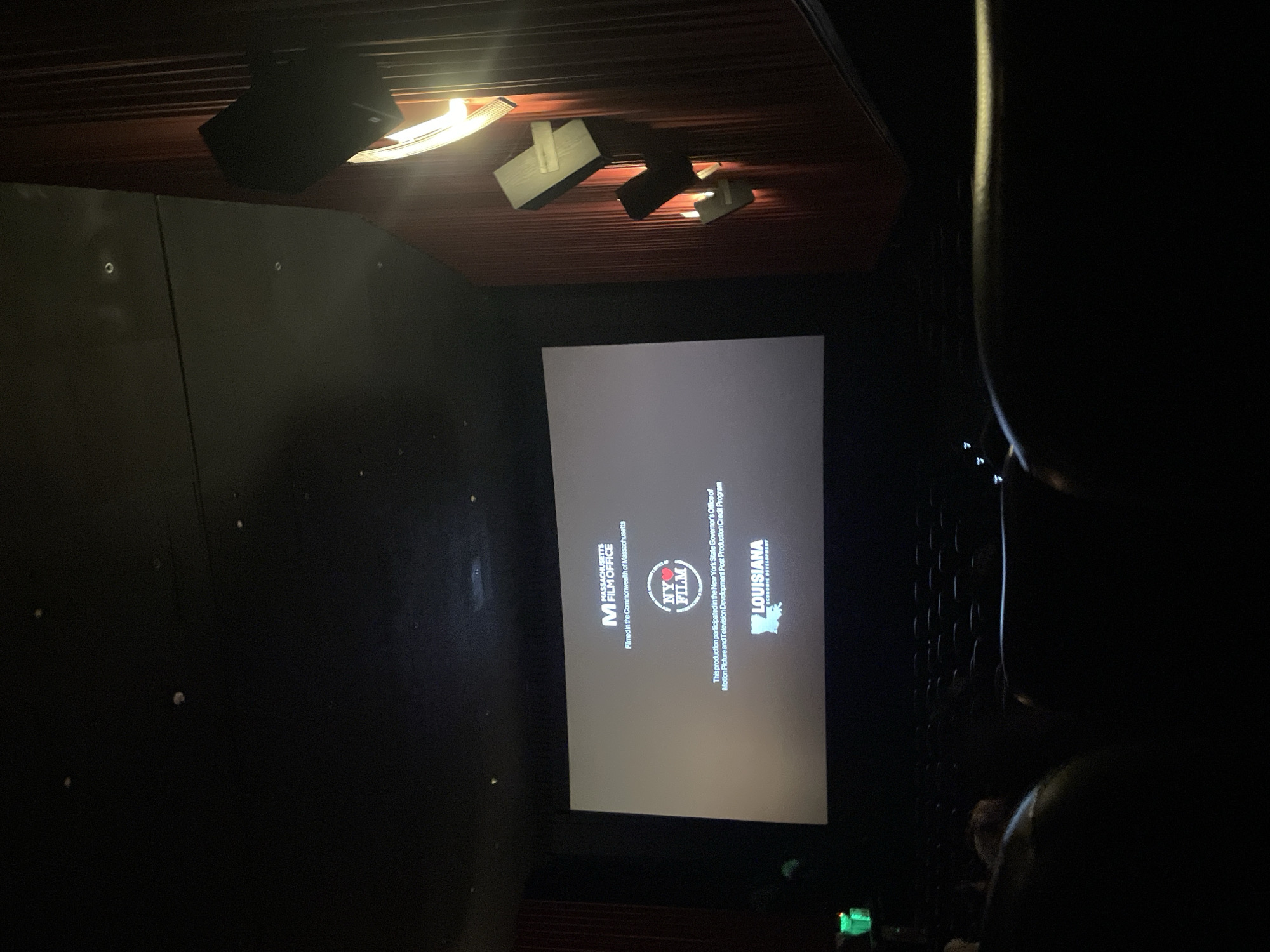The image depicts a movie theater screen displaying logos of film offices from Massachusetts, New York, and Louisiana, indicating their involvement in the movie's production. The theater's ceiling has lights and speakers. The lighting is dim, and there are blurry silhouettes of people sitting in the foreground. The location in the city of Buenos Aires, Argentina, is not visible in this image.