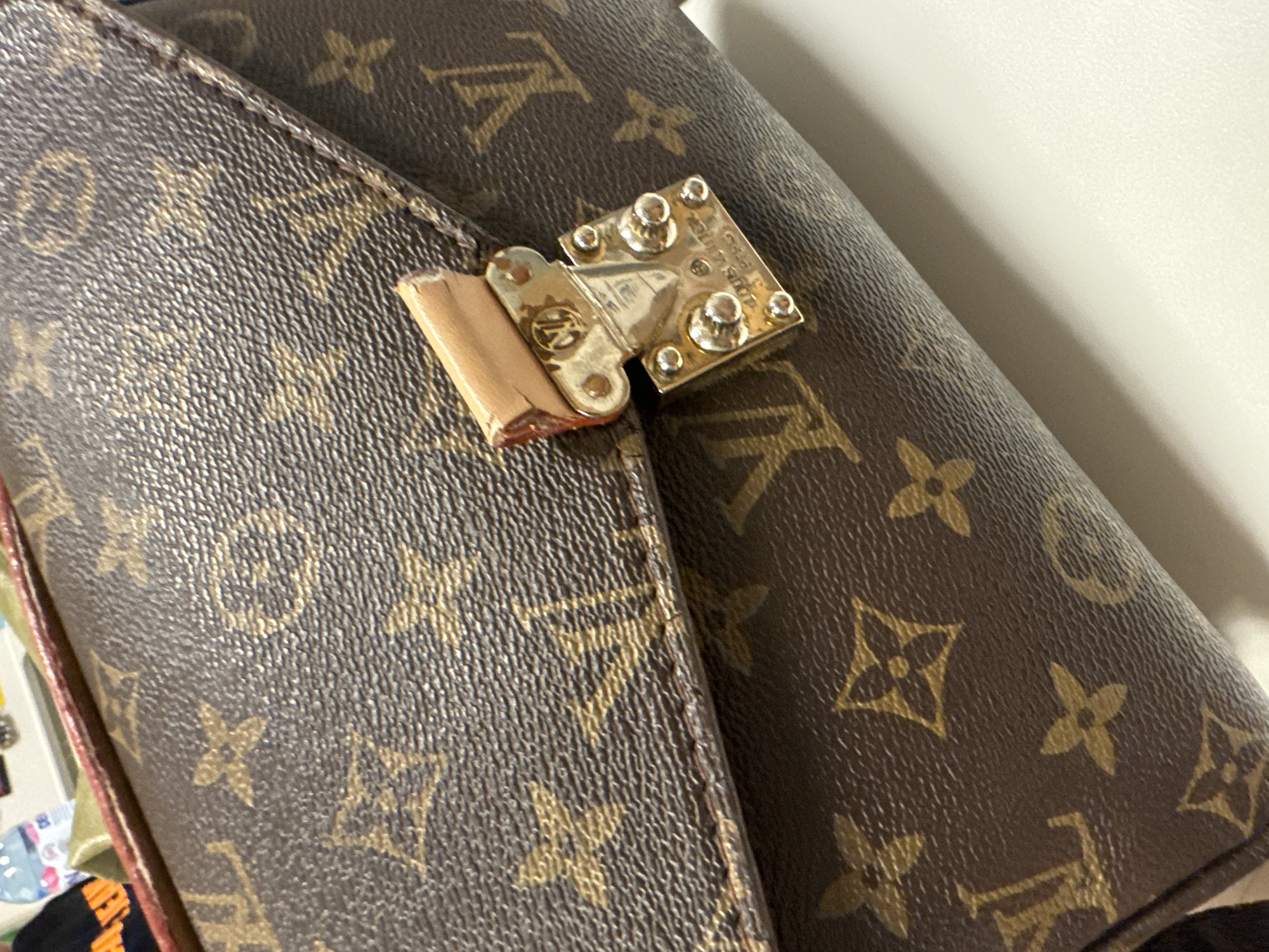 A close-up shows a Louis Vuitton handbag, with the brand's signature monogram pattern. The bag is closed by a metal clasp, which appears to be damaged. The environment suggests an indoor location. The location in the city of Unknown, Unknown cannot be determined.