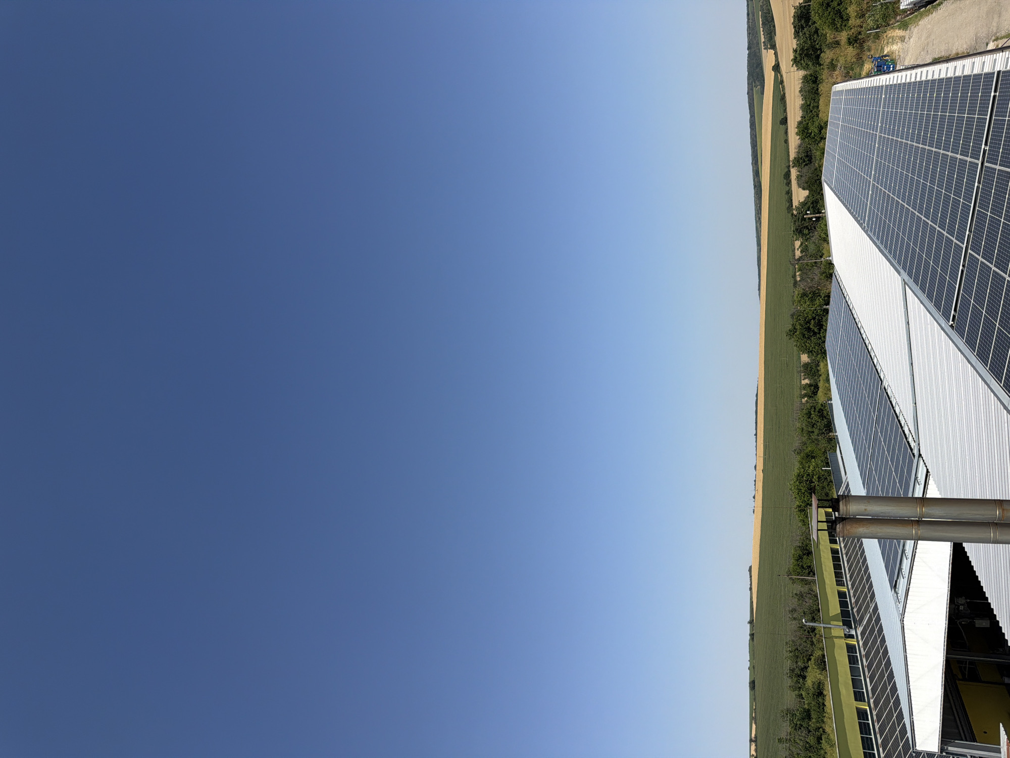 This image captures an outdoor scene with a clear blue sky dominating the upper portion. In the middle ground, a sprawling rooftop covered in an array of solar panels is visible. These panels are neatly arranged and reflect the bright sunlight. To the left, another section of a building with a yellow facade is partially visible, also featuring solar panels on its roof. In the background, a landscape unfolds with rolling hills covered in agricultural fields, showcasing a mix of green and golden-brown crops, indicative of a rural setting. Two prominent metal chimneys rise from one of the solar-panel-covered roofs. The overall scene suggests a modern facility utilizing renewable energy within a natural, agricultural environment. The location is identified as Dolno Ablanovo, Bulgaria.
