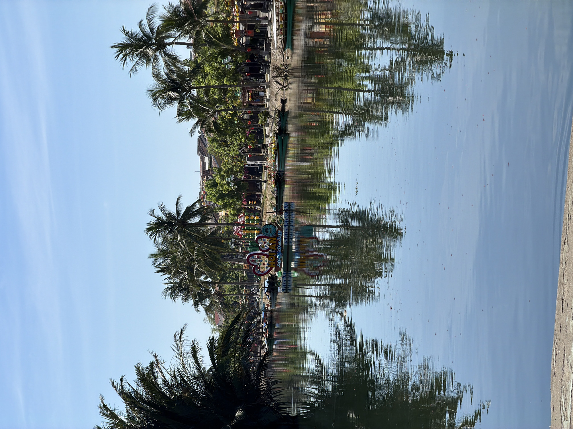 This image depicts a serene river scene in Hội An Ward, Vietnam. The water is calm, reflecting the surrounding environment, including lush palm trees, buildings with traditional architecture, and a bright blue sky. In the foreground, a pathway or embankment is visible. The river is lined with boats, some of which are traditional Vietnamese boats, and others appear to be for tourist transport. A decorative floating structure with colorful lights and heart shapes, along with signage that appears to read "HOI AN", is situated in the middle of the river. The overall atmosphere is peaceful and picturesque, suggesting a typical day in this popular tourist destination.