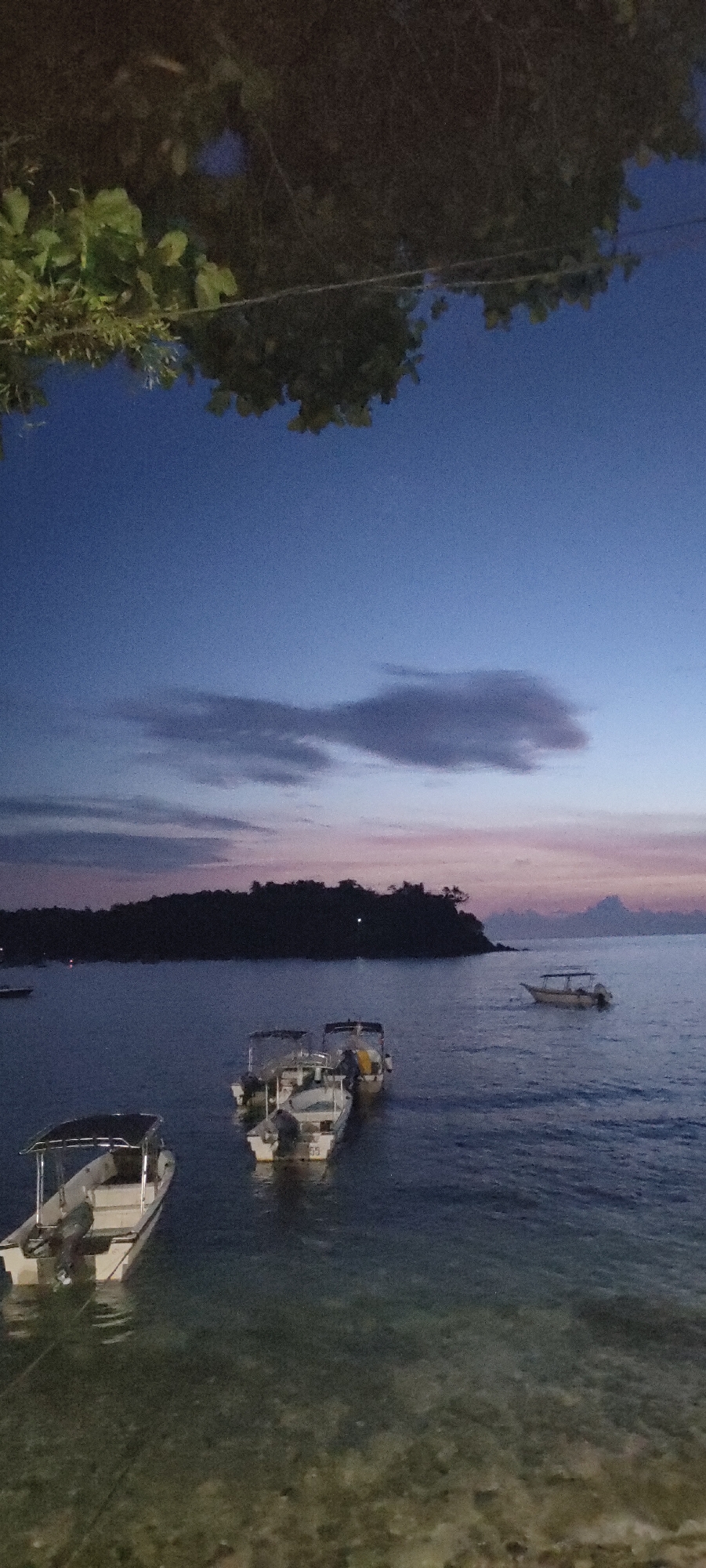 This image captures a serene evening scene at a coastal area in Sabang, Indonesia, likely near the Iboih Beach or a similar location known for its clear waters and calm bays. The time of day is twilight, with the sky transitioning from a deep blue to softer hues of purple and pink near the horizon, indicating either sunrise or sunset. A few wispy clouds are scattered across the sky.
The foreground is dominated by the shallow, clear water of the bay, revealing a sandy or rocky seabed. Several small motorboats are moored or gently drifting in the water. In the lower left, a white boat with a dark canopy is clearly visible, its engine partially submerged. Further into the bay, two more boats are clustered together, also white with canopies and visible outboard motors. In the mid-ground, a single boat is positioned further out in the water.
In the background, a dense, dark silhouette of a forested island or peninsula stretches across the horizon. The lush vegetation of the island contrasts with the open sky and water. The overall impression is one of peacefulness and natural beauty, characteristic of coastal regions in Sabang during the early evening.