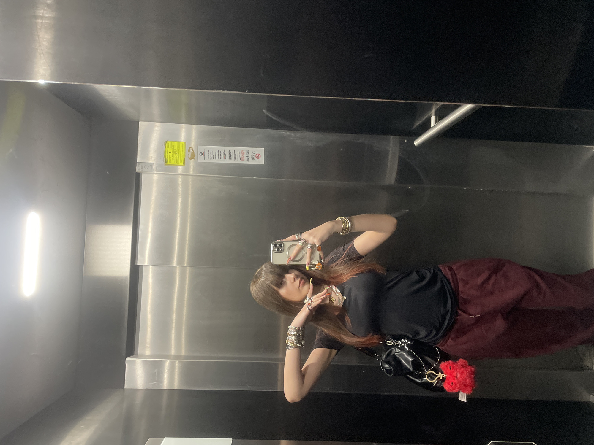 A woman is taking a selfie in an elevator. She is wearing a black shirt and maroon pants. She is holding a phone in her right hand and has several rings and bracelets on both hands. She has long brown hair with some red highlights. There is a black purse with a red flower charm hanging from it near her left hip. The elevator is stainless steel and has a handrail. There is a notice in both English and Hebrew on the wall of the elevator. The specific location in Raanana, Israel is unknown.