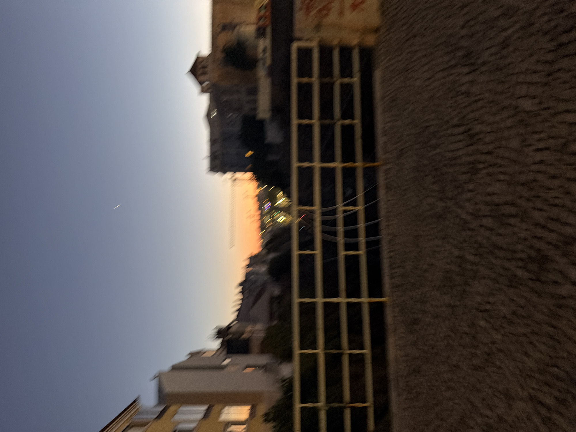 The image shows a view of buildings and the sky in Cascais, Portugal. The orientation is rotated, with the sky occupying the left side and buildings on the right. The sky is a gradient of blue to a warm orange, suggesting it is either sunrise or sunset. A thin white line, possibly an airplane contrail, is visible in the sky.
On the right side of the image, there are several buildings. One building appears to be made of rough, textured material, possibly stone or stucco. Adjacent to this is a structure with a grid-like pattern of light-colored bars, possibly a window grate or a ventilation shaft. Further to the left, more buildings are visible, some with visible windows and architectural details. The lighting suggests it's dusk or dawn, with artificial lights starting to appear or fade in the buildings. The overall scene depicts an urban environment under a twilight sky. There is no visible text in the image.