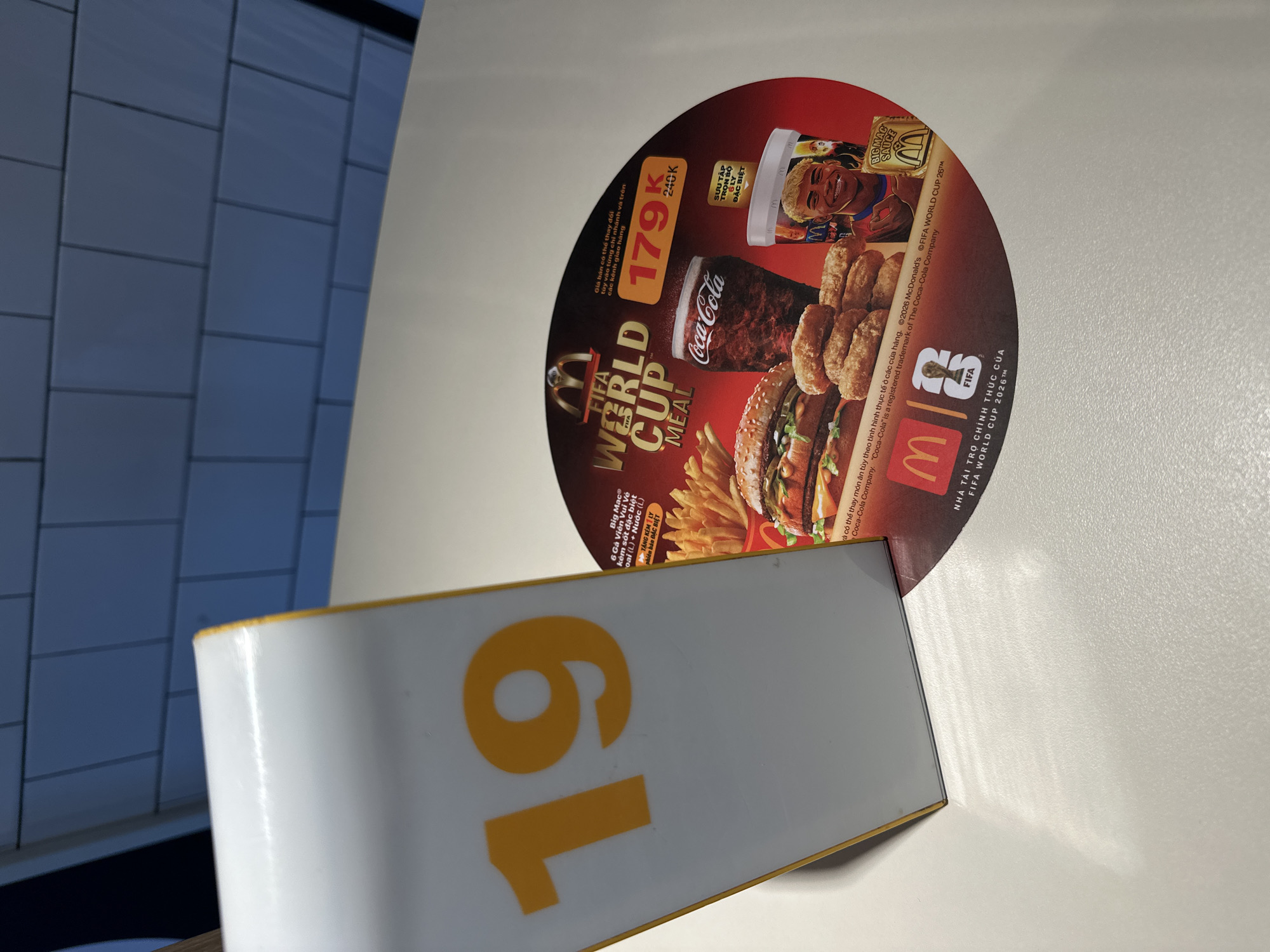 The image shows a promotional circular advertisement from McDonald's for a "FIFA World Cup Meal" deal. The advertisement features a burger, fries, a Coca-Cola drink, and a collectible cup with a soccer player on it. The text indicates the meal costs 179,000 VND and includes a special sauce. The advertisement is likely displayed in a McDonald's restaurant, possibly as a table tent or a circular display. The overall impression is of a fast-food promotion tied to a major sporting event. The "19" on the table tent suggests this might be table number 19. The background shows a tiled wall and a white table, typical of a restaurant setting. The lighting suggests it could be daytime. The advertisement mentions "Nha Trang Ward, Vietnam" in the text.