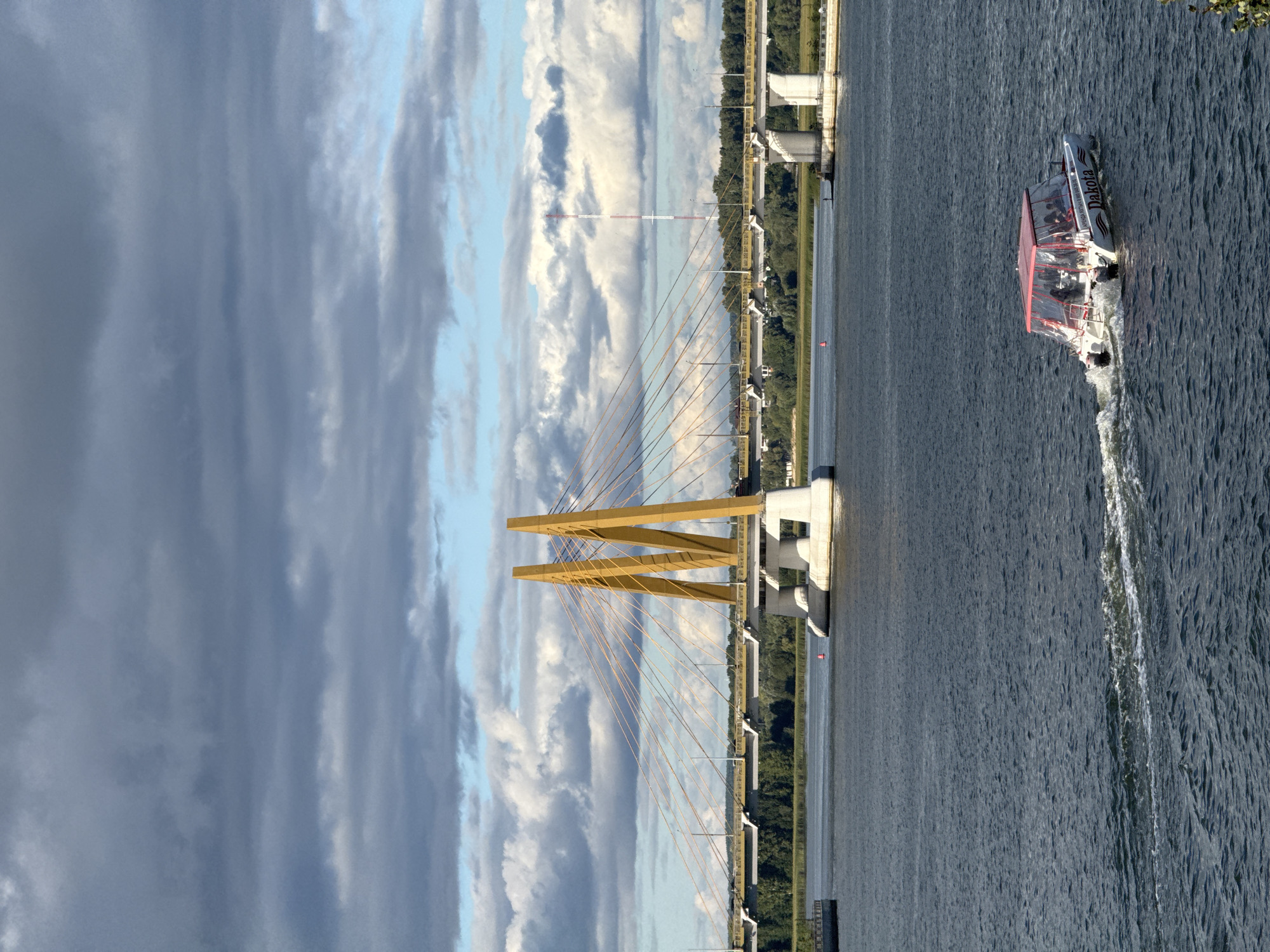 The image depicts a wide river with a modern cable-stayed bridge in the background. The bridge features two prominent yellow pylons supporting a network of cables, with a road deck spanning the river. A boat with a red canopy, labeled "Dakota," is moving across the water in the foreground, leaving a wake. The riverbanks are lined with trees, and a distinctive red and white striped tower is visible in the distance beyond the bridge. The sky is partly cloudy with dramatic, dark clouds interspersed with patches of blue. This scene is the Kazansky (Kazan) Bridge over the Kazanka River in Kazan, Russia.