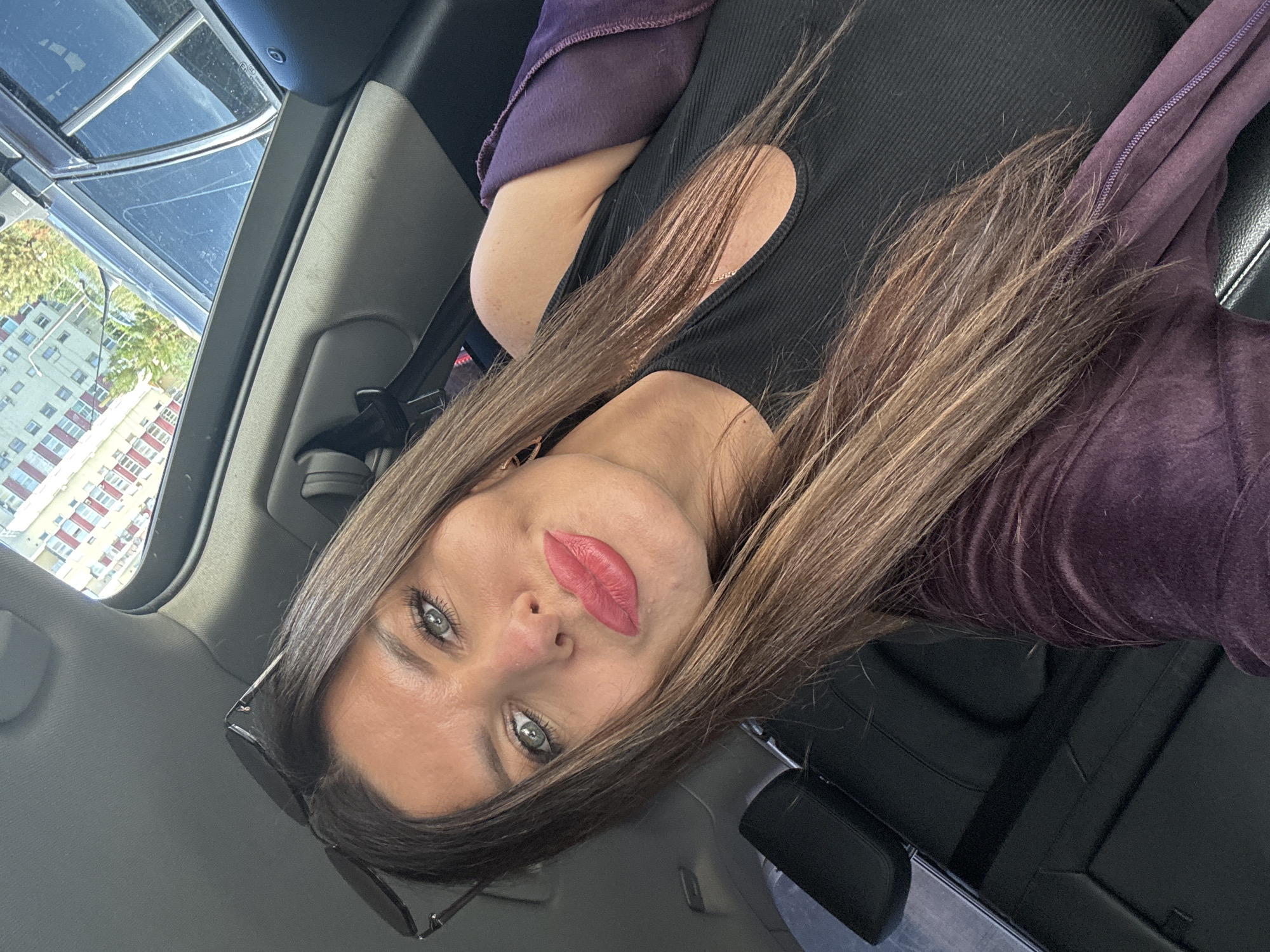 The image shows a woman sitting in a car, with the camera angled upwards. She has long, brown hair and is wearing a black top with a cut-out detail on the shoulder, and a purple hooded jacket. Her lips are painted with a bright pink lipstick, and she has green eyes. Sunglasses are resting on her lap. Through the car's sunroof, an apartment building with red and white accents is visible, along with trees and a bright sky, suggesting it is daytime and the weather is clear. The interior of the car, including black leather seats and a seatbelt, is also visible. The scene appears to be in Chaikovsky, Russia. The woman is looking directly at the camera, with a neutral expression.