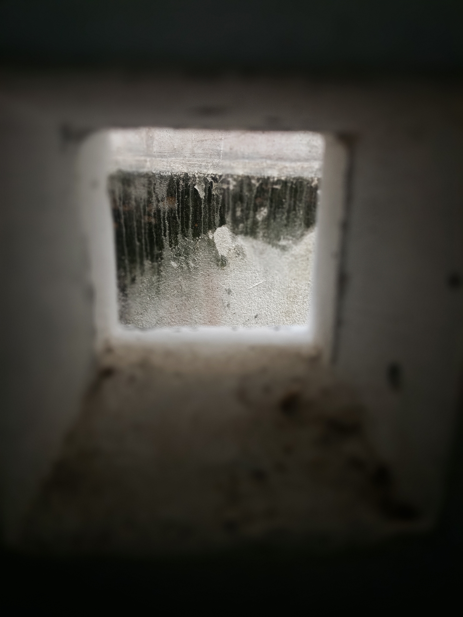 The image shows a square opening in a weathered, possibly concrete, structure. The view through the opening is obscured by what appears to be dirty or frosted glass, or possibly a buildup of grime and dripping condensation. Vertical streaks are visible on the glass. The area immediately surrounding the opening is also aged and potentially stained. The blurred foreground indicates the viewpoint is from within a confined space looking outward. The location in Bireuen, Indonesia, is not apparent from the image itself.