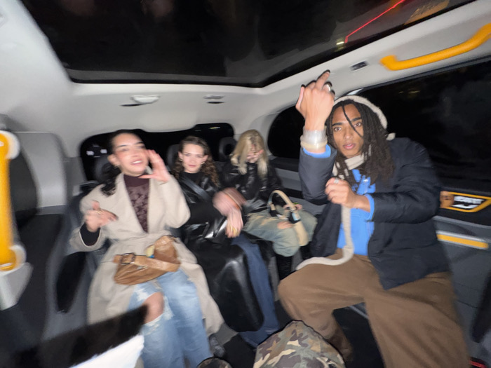Inside a taxi at night, four young adults are captured. The person on the right, sporting dreadlocks and a beanie, boldly raises a middle finger. The two figures in the middle exhibit smiles. The person seated on the left is gesturing with both hands, seeming animated. All are dressed stylishly, each with a unique bag. The taxi's interior is visible, complete with a clear roof panel and a yellow safety handle. The image suggests a jovial atmosphere during an evening outing.