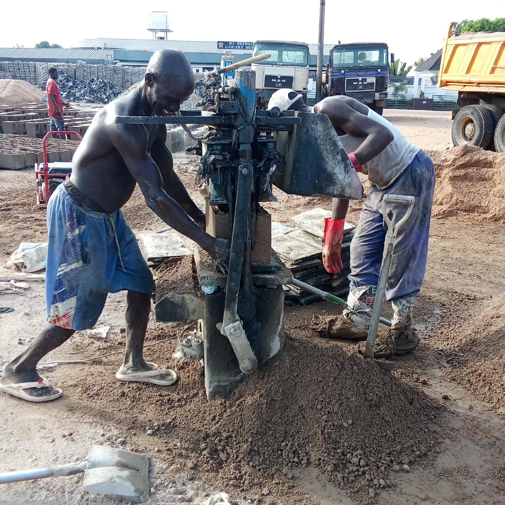The image depicts an outdoor, industrial scene in Akwanga, Nigeria, centered around manual block-making. In the foreground, two men are operating a hand-powered block-making machine. The man on the left, bare-chested and wearing blue shorts and flip-flops, is actively pulling the machine's lever. He appears focused, with a slight smile. The man on the right, wearing a white tank top, blue trousers, and work boots with red gloves, is pouring raw material from a scoop into the machine. The ground around them is a mix of dirt and sand, with mounds of earth and traces of wet concrete. A metal shovel rests on the ground nearby.
In the mid-ground, a third man in a red shirt and jeans stands amidst large stacks of newly produced concrete blocks, some still drying. A red hand truck or cart is also visible in this area. The background features several large vehicles, including two "M.A.N" branded trucks and a yellow dump truck. Behind the trucks, low-rise industrial buildings with metal roofs can be seen. One building displays partial text, "RI-SEINGO" and "LUXURY HO", likely part of a business name. The clear sky indicates a bright, sunny day, highlighting the laborious nature of the work. The scene captures a typical manual construction materials production process in a busy, functional setting.