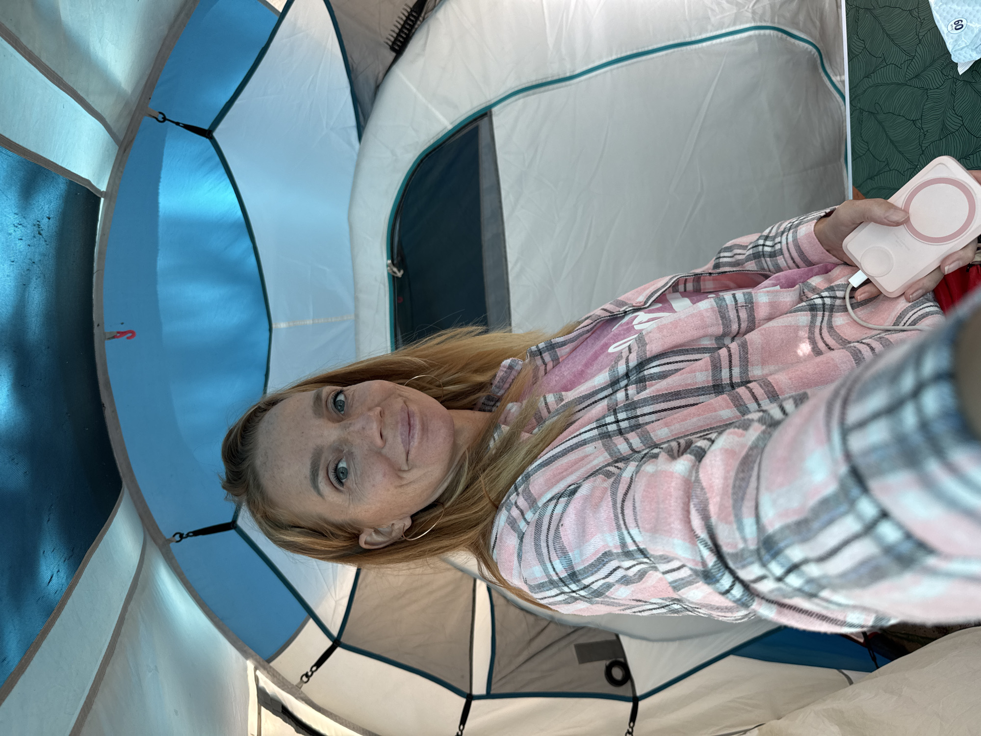 In the city of Zelenaya Roshcha, Russia, a woman is pictured inside a tent. She has blonde hair and is wearing a pink and white plaid shirt. She is holding a pink portable charger in her right hand. The tent is blue and white, with a window on the right side.