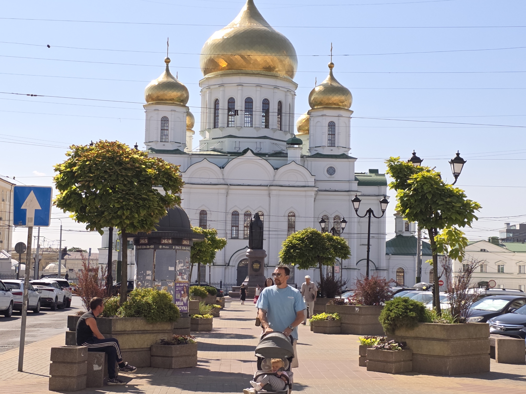 The image shows a sunny day scene in front of the Rostov Annunciation Cathedral in Rostov-on-Don, Russia. A man in a blue t-shirt is pushing a stroller with a baby inside. A man in a black tank top and track pants is sitting on a concrete planter to the left. In the background, the white cathedral with golden domes is visible, along with trees, streetlights, and cars parked on the street. The foreground features a paved walkway lined with concrete planters filled with various plants.