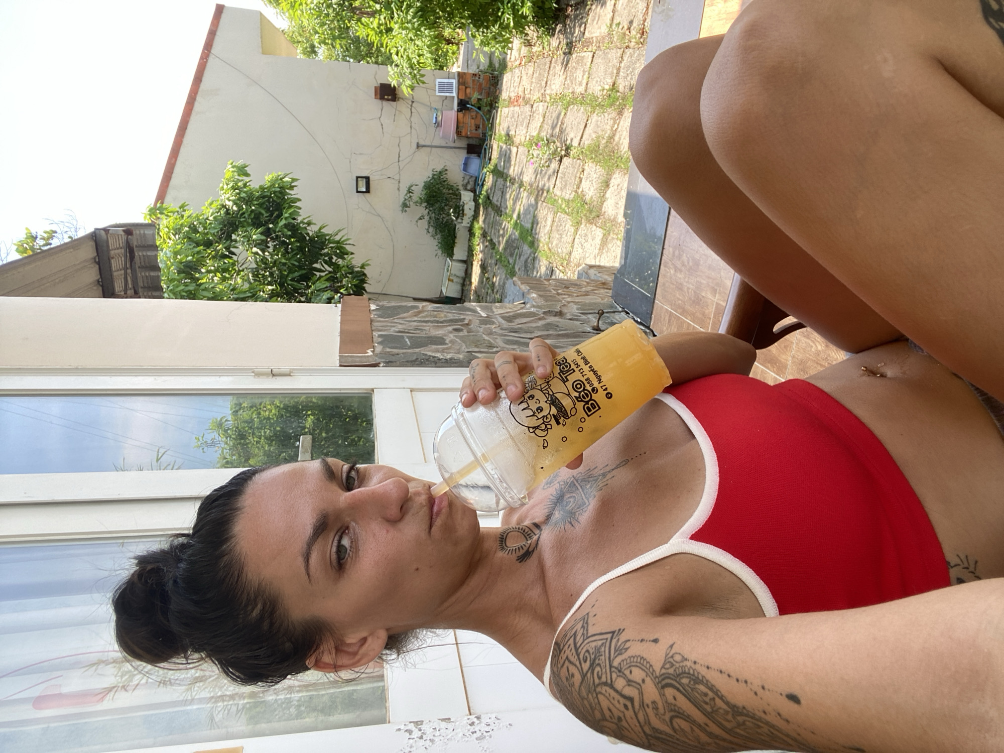 A woman with dark hair pulled up in a bun, wearing a red tank top with white trim, sits outdoors and drinks from a clear plastic cup filled with a yellow beverage. She has tattoos on her left arm, chest, and neck. The woman is positioned on a wooden chair on a tiled patio, with a stone wall and greenery in the background. The scene appears to be in Mũi Né, Vietnam, given the presence of tropical vegetation and architecture commonly found in the area.
