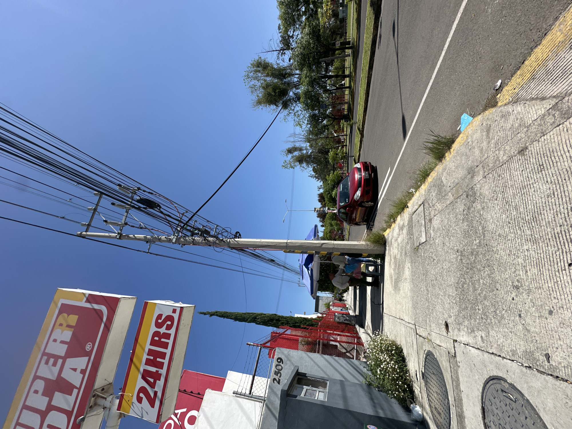 The image shows a street scene in San Bernardino Tlaxcalancingo, Mexico. On the left, a gray building with a window and the number "2509" painted on it is visible. Above the building, two signs are mounted on a pole; the top sign reads "SUPER" in red letters on a yellow background, and the bottom sign reads "24HRS." in red letters on a yellow background. A sidewalk runs along the building, with a street to the right. There are some white flowers growing near the building. A power pole with many wires stands on the sidewalk. Two people are standing under a blue umbrella next to a smaller blue umbrella. A red car is driving on the street. The sky is clear and blue, and there are trees and other buildings in the background.