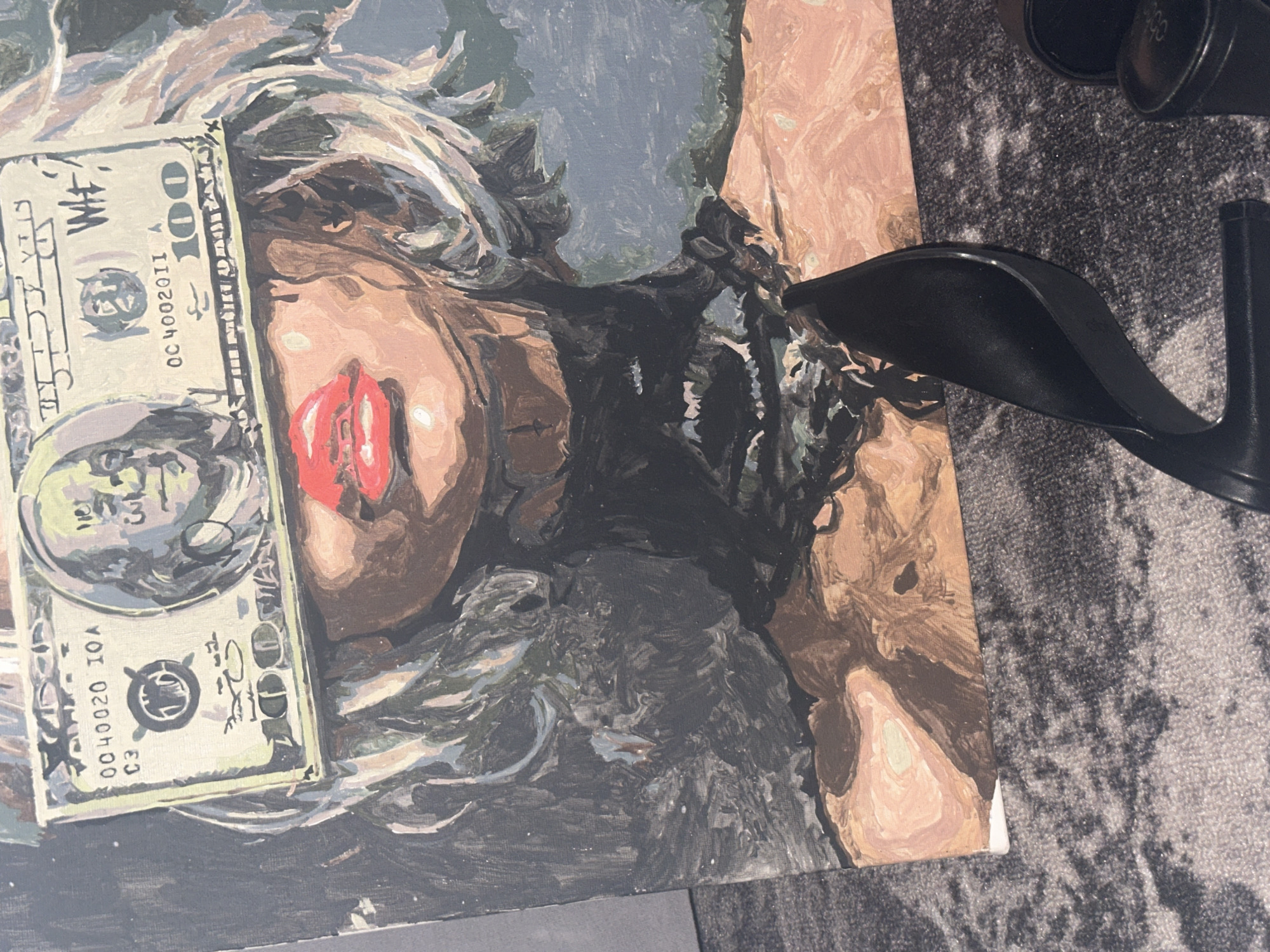 The image shows an artwork of a woman's face, partially obscured by a one hundred U.S. dollar bill. The artwork is rendered in a paint-by-numbers style with visible brushstrokes and muted colors. The woman has red lips, fair skin, and fair hair. A black, high-heeled shoe is resting on top of the artwork. The surface beneath is a carpet with a grey, textured pattern. The location cannot be identified based on the provided image.