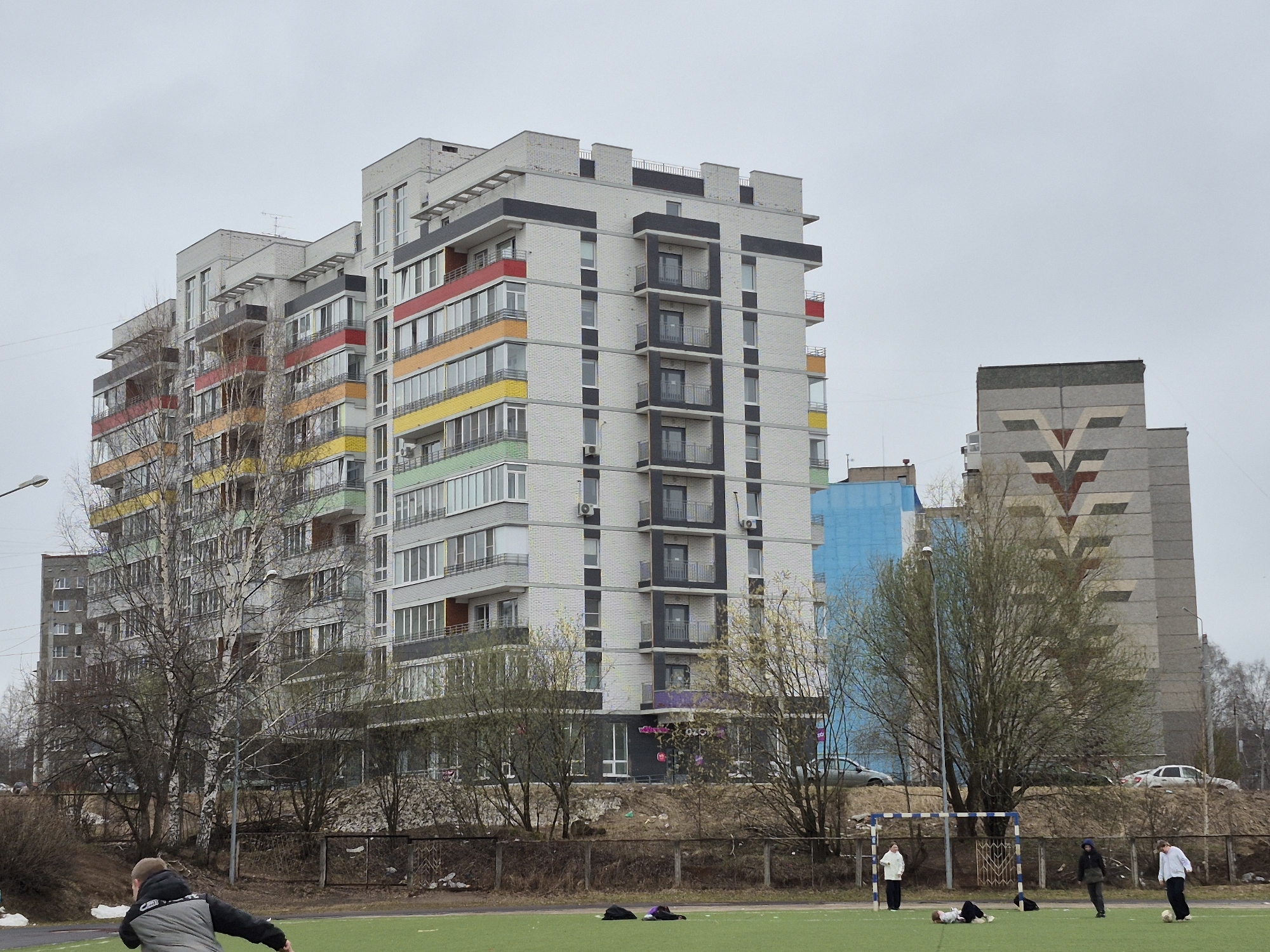 The image captures an urban scene in Chaikovsky, Russia, featuring multiple residential buildings and a green artificial turf field under an overcast sky.
The most prominent structure is a multi-story, modern apartment building with a light-colored brick facade. Its balconies are adorned with distinct horizontal bands of color, progressing from red at the top to orange, yellow, and light green further down, creating a rainbow-like gradient. Dark gray vertical and horizontal elements, along with smaller dark-railed balconies, punctuate the building's design. The ground floor of this building features a retail space with large dark windows, where the purple and white logo for "OZON" (a Russian e-commerce company) is clearly visible. To the right of this building, a taller, plainer gray residential block stands, distinguished by a large, geometric pattern across its upper facade, composed of shades of gray, white, and reddish-brown, resembling an abstract 'V' or 'Y' motif. A smaller, partially visible blue building can be seen between and behind the trees.
In the foreground, a vibrant green artificial turf field is present, separated from the buildings by a fence and a slight incline of bare earth. Several individuals are scattered across the field. In the bottom left, a person in a black and gray jacket is captured in mid-movement. Towards the center, two dark bags or jackets are left on the grass. Further right, a soccer goalpost with blue and white stripes stands. Around the goal, three individuals are visible: one person in a white top stands casually, another in a dark jacket walks, and a third person in a white top is near a soccer ball. Another individual is lying down on the field near the goal. The trees bordering the field are mostly bare, though some show hints of budding leaves, suggesting early spring.
The time of day is daytime, but the uniformly gray sky indicates overcast weather. The scene conveys a casual, everyday atmosphere with people engaged in outdoor activity or relaxation in a residential setting.