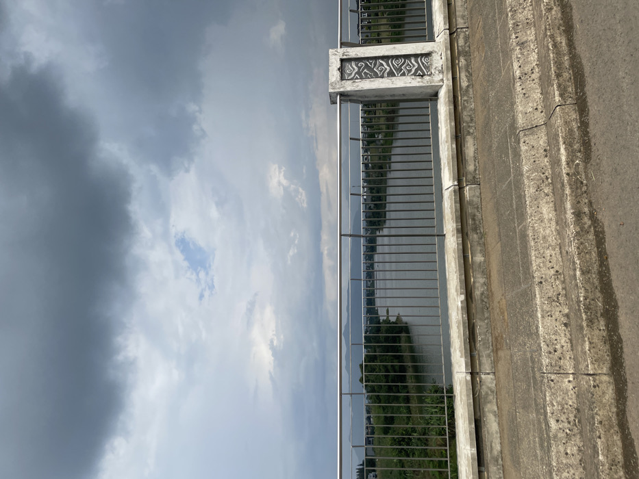 A metal railing separates the viewer from a river that is flowing in the distance, surrounded by a low-lying area of green vegetation on either side. The river is framed by a distant mountain range, and the sky is overcast with gray clouds. The scene is captured from a concrete bridge, with a concrete sidewalk in the foreground and an asphalt roadway just beyond the edge of the frame. There is a concrete pillar with an ornate design on its surface to the right of the railing. The location is a bridge in Padalarang, Indonesia, at latitude -6.865 and longitude 107.461.