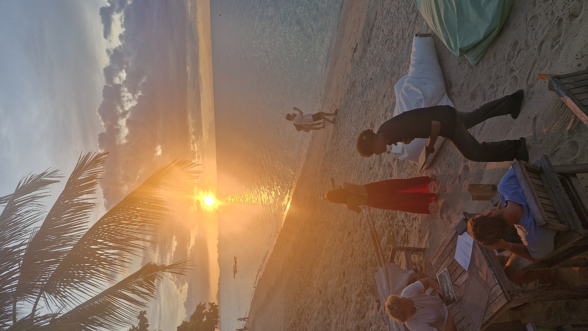 The image depicts a beach scene at sunset in Baan Thong Po, Thailand, characterized by warm, golden light filtering through palm fronds. Several individuals are present on the sandy shore. In the foreground, a man with curly hair wearing a dark shirt and pants walks, holding a thin object. To his left, a person with blond hair sits at a wooden table with papers, wearing a white t-shirt. Another person, viewed from behind, sits in a wooden chair wearing a blue t-shirt.
Further down the beach, a woman in a red dress stands near a person in a white shirt and dark shorts, who appears to be walking away from the viewer. In the distance, more figures are visible walking along the shoreline, and a few small boats are moored on the calm sea. The sky is filled with clouds tinged with orange and yellow from the setting sun, which casts a shimmering reflection on the water. Several large, soft seating items, resembling beanbags, are scattered on the sand, one white and one light green. The overall atmosphere is relaxed and tranquil, typical of a tropical beach at dusk.
