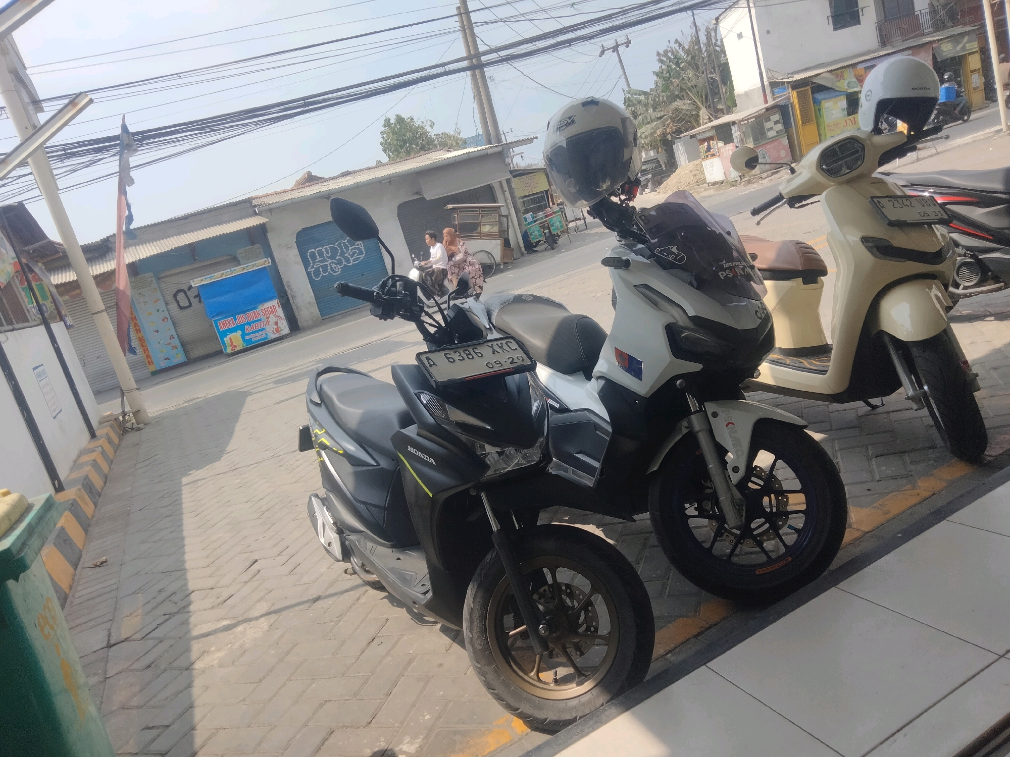 In Pisangan Jaya, Indonesia, two motorcycles are parked on a brick-paved street. The motorcycle on the left is black and gray, with a black windshield and a yellow stripe on its side panel. Its license plate reads "A 6386 XKC 09-29". The motorcycle on the right is cream-colored, with a brown seat and a white helmet resting on its handlebars. Its license plate reads "A 2342 UBD 05-31". A third motorcycle, partially visible behind the cream-colored one, is dark-colored with red accents. In the background, there are several buildings with corrugated metal roofs and closed storefronts, some with colorful advertisements and graffiti. A few trees are visible in the distance. Overhead, a tangle of power lines crosses the sky. A woman in a headscarf and a man are seated on a motorcycle in the middle ground.