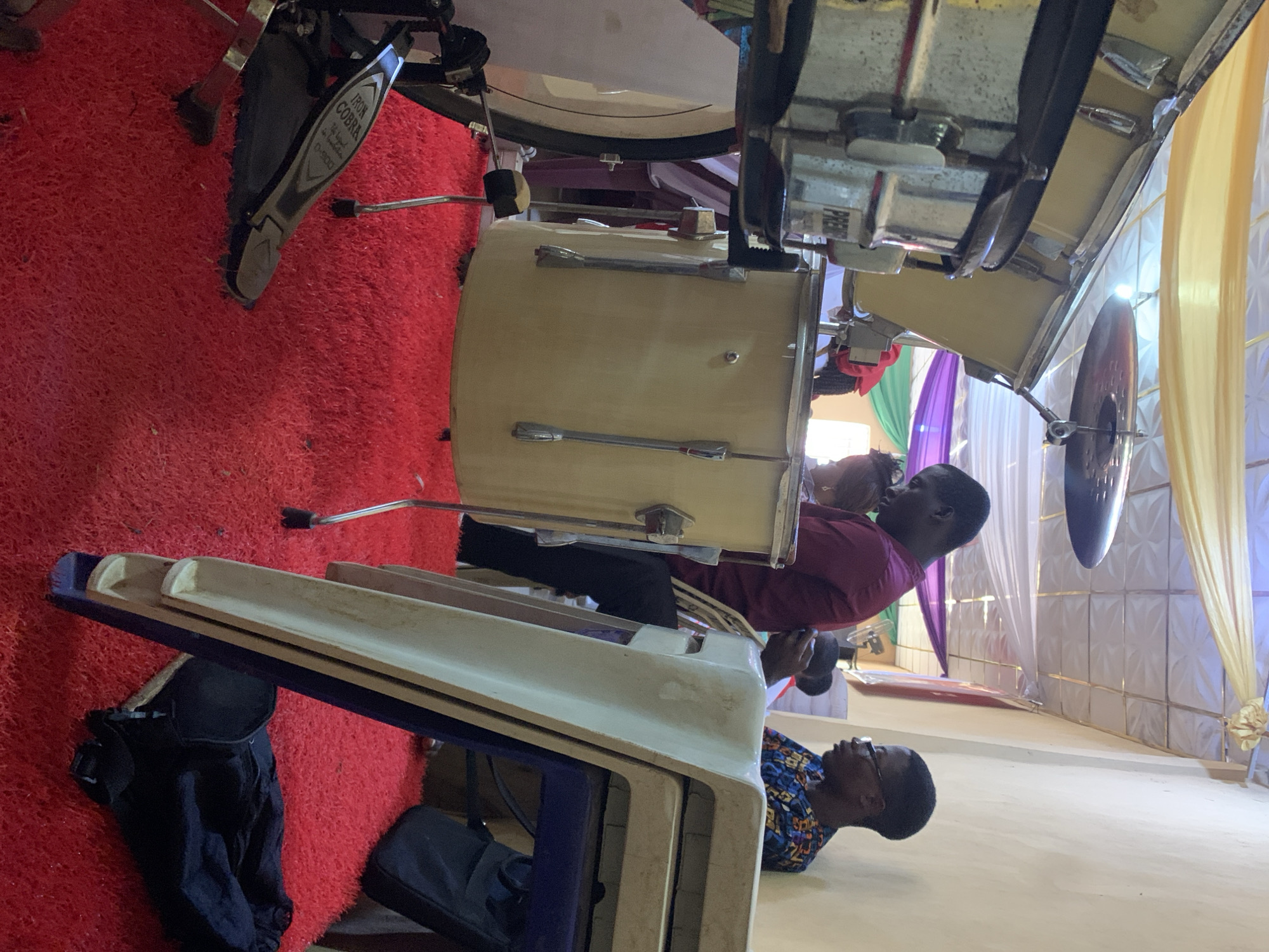 The scene appears to be indoors in the city of Akpaka Forest Reserve, Nigeria. A set of drums is prominently featured, positioned on a red carpet. A man wearing a maroon shirt sits near the drums. Another man wearing glasses and a patterned blue shirt is situated closer to the floor, near what appears to be stacked seating. The backdrop includes white and gold draped fabric.