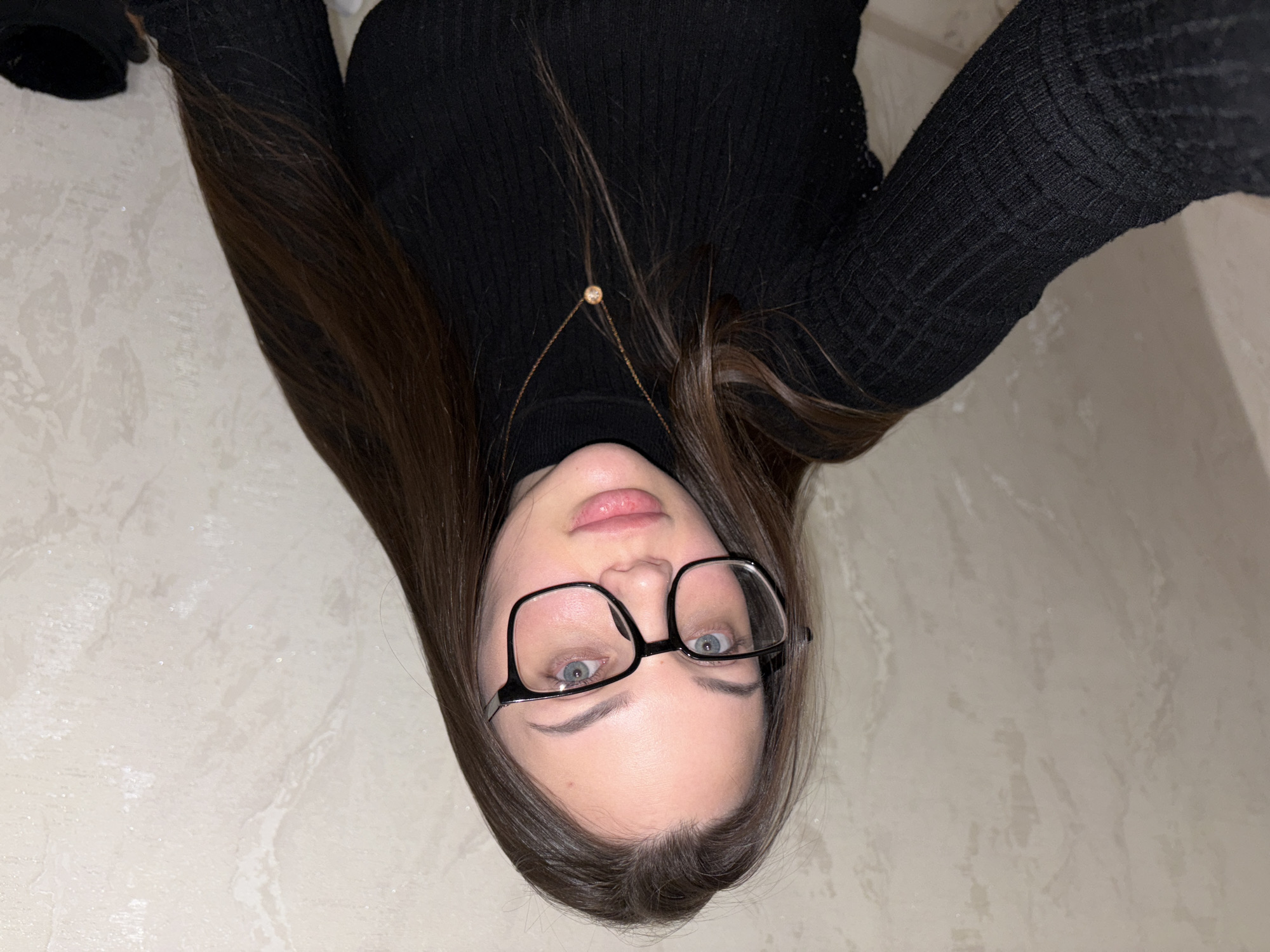 A young woman is pictured from an upside-down perspective in a close-up selfie. She is wearing a black turtleneck sweater and eyeglasses with a black frame. A delicate gold necklace is visible around her neck. She has fair skin, light blue eyes, and her dark brown hair is long and straight. The background is a light-colored wall with a subtle, textured pattern. The location is not discernible from the image, and there is no indication that the picture was taken in Chaikovsky, Russia.