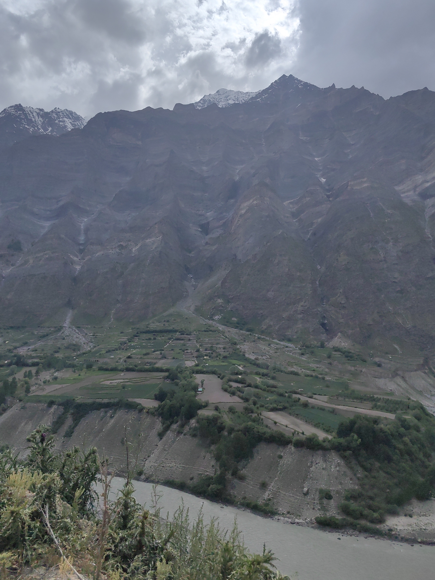 The image depicts a vast mountainous landscape near Tandi, India. In the foreground, lush green vegetation and thorny bushes are visible, with a winding river flowing to the right. The middle ground features terraced agricultural fields and scattered trees, suggesting a rural settlement. The background is dominated by immense, rugged mountains with a cloudy sky above, hinting at a high-altitude, remote location. The overall scene conveys a sense of natural grandeur and the peaceful coexistence of human activity with the environment.