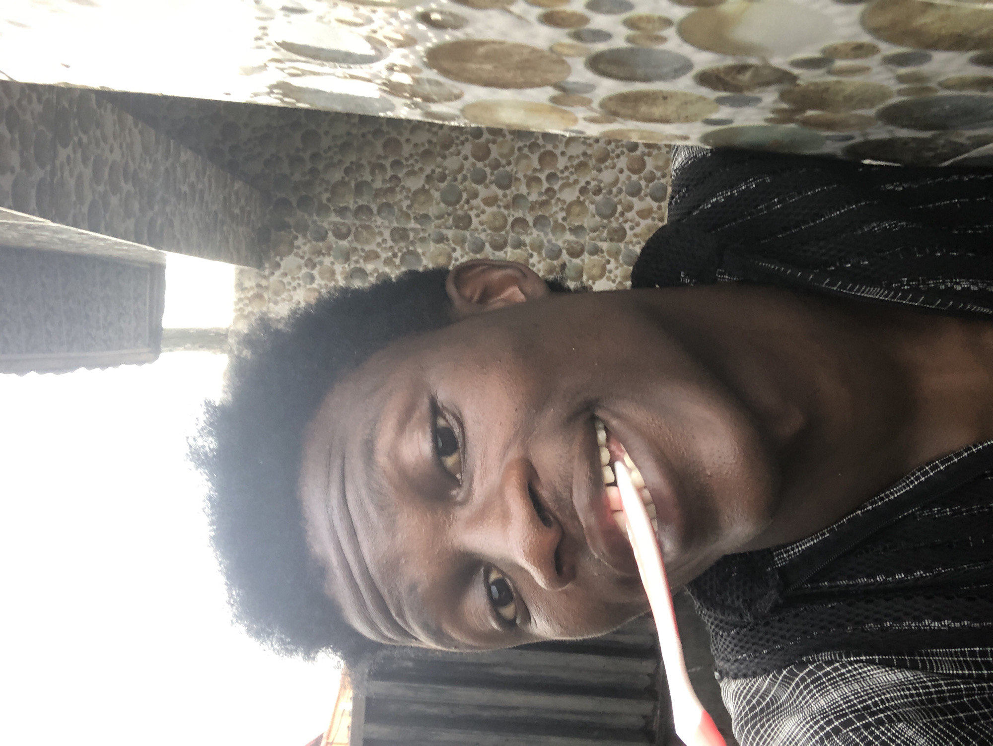 A young Black male is captured in a close-up, selfie-style photograph, brushing his teeth outdoors. He is wearing a black and white checkered shirt with a mesh overlay. A pink toothbrush is in his mouth, actively being used. The background suggests an urban environment in Ado, Nigeria, with a corrugated metal fence visible to the left and a textured tiled wall to the right. The lighting is bright, indicating daylight.