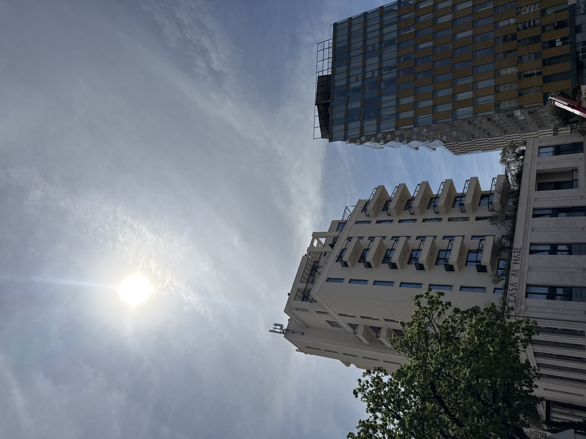 The image, taken from a low angle, captures the upper portions of two buildings against a backdrop of a bright sky with scattered clouds. The building on the left is predominantly white with multiple balconies and the inscription "CASA AL MARE" on its side. The building on the right is a modern structure with a grid-like facade of alternating gold and glass panels. A green tree is partially visible near the base of the white building. The sun is visible in the top left quadrant, creating a lens flare. Location is Unknown, Unknown.