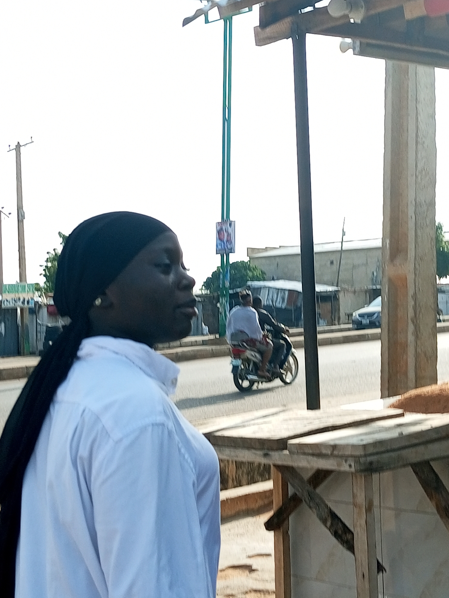 An overhead shot captures a young Black woman with a black hijab and a white collared shirt standing in profile. She is facing left, and her right earlobe is adorned with a small pearl earring. Behind her, a street scene unfolds with a motorcycle carrying two people traveling away from the camera. The driver wears a dark shirt, and the passenger, facing forward, is in a white shirt. A silver sedan is parked on the side of the road further down, with a few buildings and trees visible in the background. In the foreground, a wooden structure made of planks and support beams is partially visible, with a textured brown material resting on its surface. The scene appears to be outdoors, under a bright sky, suggesting an urban environment in Tempure, Nigeria.