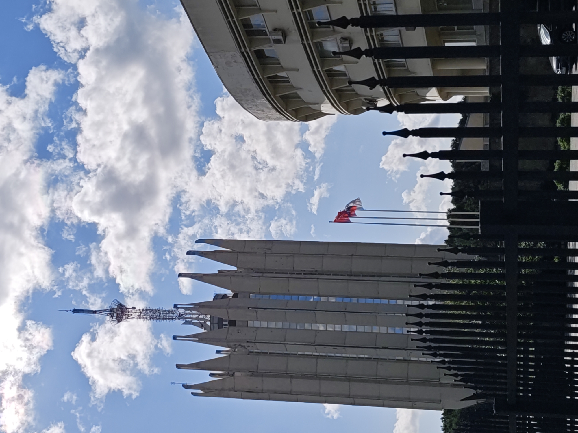 The image captures a scene in Saint Petersburg, Russia, dominated by two large buildings and a black metal fence in the foreground. The building on the left is a tall, angular tower with a distinctive pointed top, reminiscent of a spire or crown, and a lattice radio tower extends from its apex. The building on the right is a curved, light-colored structure with multiple floors and evenly spaced windows, some of which have visible air conditioning units. Two flags, one red and one white and red, are visible flying on flagpoles between the two buildings. The sky is blue with scattered white clouds. The fence in the foreground is elaborate, with pointed finials atop each vertical bar. A dark-colored car is partially visible behind the fence on the right. The overall impression is of a formal or governmental area.