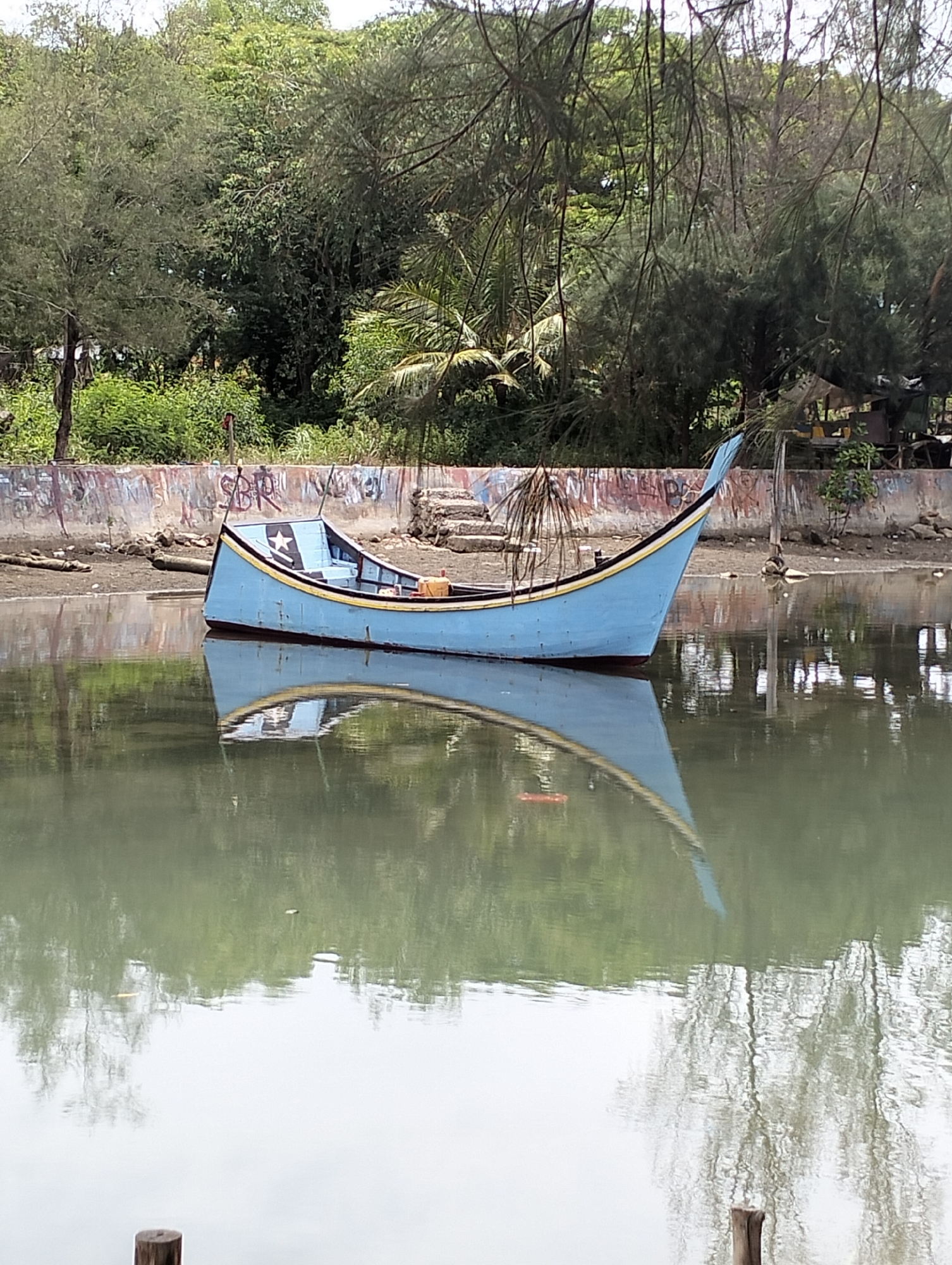 The image shows a light blue boat with black and yellow trim, featuring a black star on its side, floating on calm water that reflects the boat and surrounding trees. The boat is near a concrete wall with graffiti. Beyond the wall, lush green trees and vegetation are visible. Two wooden posts stand in the foreground. The location is Banda Aceh, Indonesia.