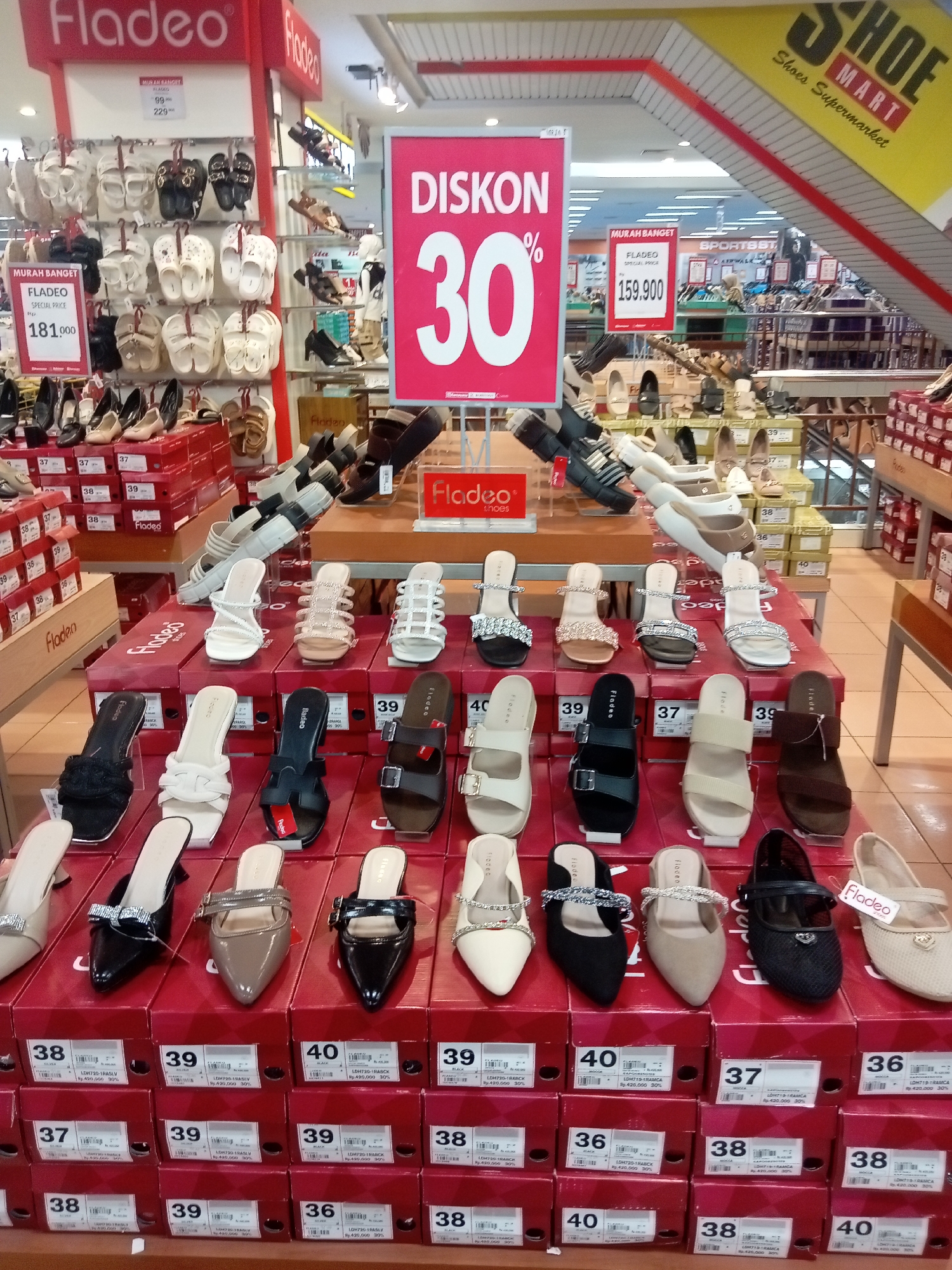 This image depicts a retail store in Batam, Indonesia, specifically a shoe department, likely within a larger supermarket or department store. The prominent branding "Fladeo" and "SHOESMART Shoes Supermarket" suggests a chain or a specialized shoe retailer. The scene is dominated by displays of women's footwear, arranged on tiered shelves and atop stacks of shoeboxes. A large pink promotional sign advertises a "DISCOUNT 30%" and shows the price "30%" in large numbers, indicating a sale event. Various styles of sandals, heels, and flats are visible, predominantly in neutral colors like black, white, beige, and brown, with some embellished with glitter or bows. Shoeboxes are stacked in neat rows, labeled with sizes and potentially product codes, with some displaying prices and discount percentages. The environment appears to be indoors, with artificial lighting illuminating the merchandise. The overall impression is a well-organized retail space focused on selling women's shoes, with an emphasis on promotional pricing.