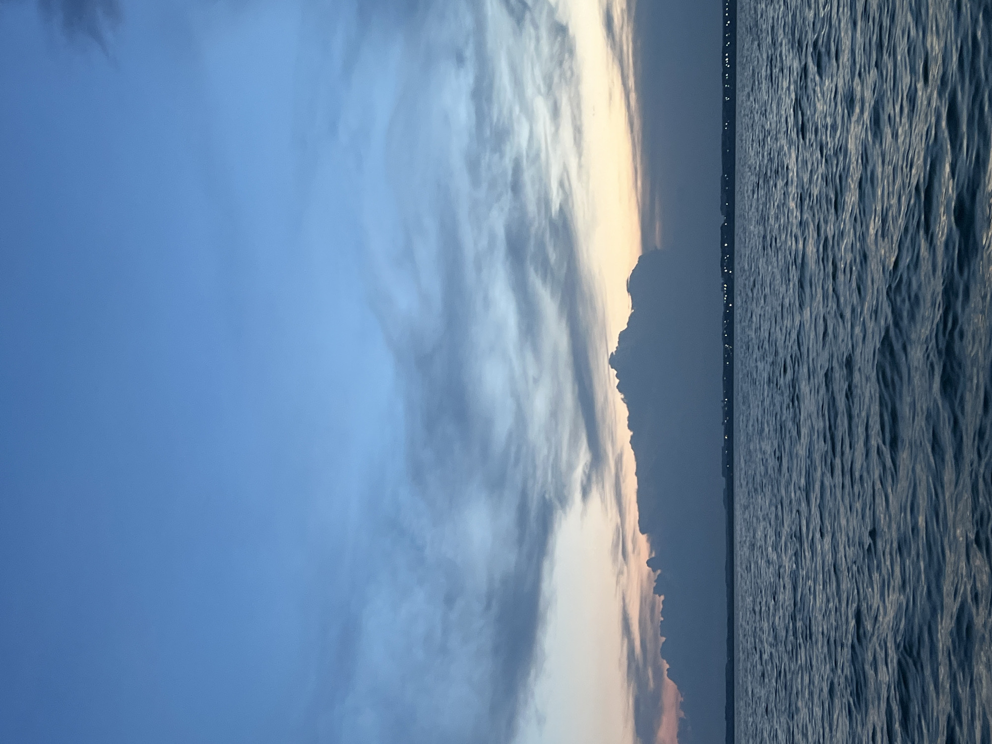 The image depicts a serene body of water under a twilight sky, with a distant shoreline. The water is a deep blue, reflecting the colors of the sky and showing gentle ripples. The sky transitions from a pale orange and pink near the horizon to a deeper blue overhead. A line of clouds, dark and textured, stretches across the middle of the image, partially obscuring the horizon. On the distant shoreline, faint lights are visible, suggesting a settlement or town. There are no people or discernible objects in the foreground or middle ground. The overall impression is one of calm and quiet. The location in the city of Novopichugovo, Russia, is not visually evident from the image itself, as it shows a natural landscape.