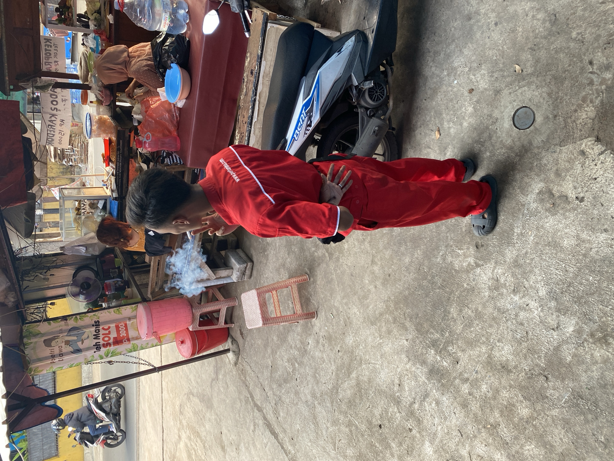 The image depicts an outdoor scene at a street-side food stall in Tangerang, Indonesia. In the foreground, a young individual wearing a red uniform with white trim stands facing away from the camera, with their back to the stall. They are smoking, and a visible plume of smoke rises from their cigarette. The individual is wearing dark-colored slides and has their hands clasped behind their back.
Behind the individual, the stall is set up under a canopy. Various food items and drinks are displayed on shelves and tables. A woman in a headscarf is visible behind the counter, interacting with the food. Several plastic stools are arranged around the stall, and a pink trash bin is present. A scooter is parked near the stall, with its rear wheel visible. In the background, a person is riding a scooter on the street, and buildings are visible. The ground is paved with concrete. The overall scene suggests a busy, everyday environment typical of street vendors in the region.