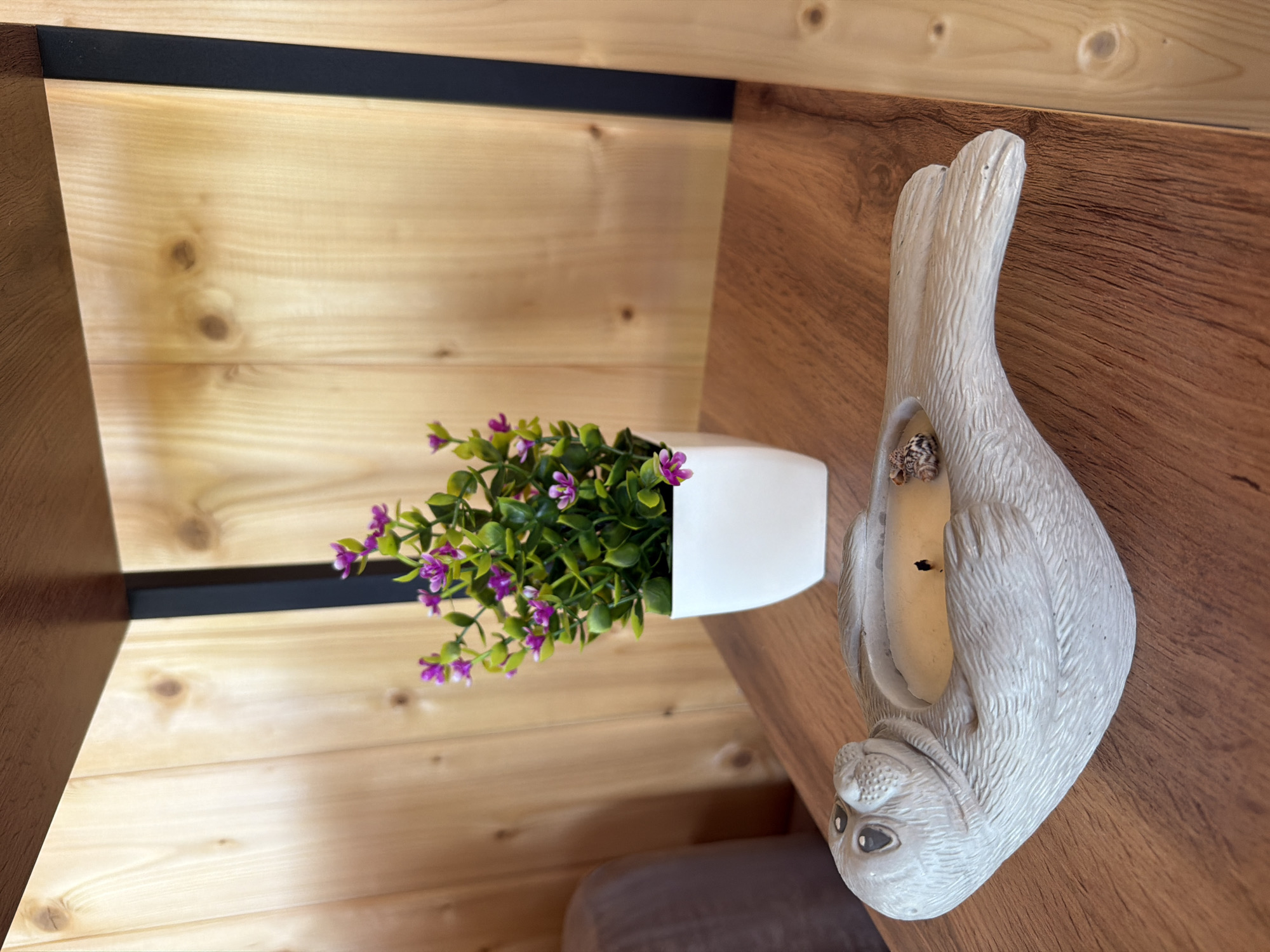 The image shows a close-up of a wooden shelf with two decorative items. On the left is a grey, seal-shaped candle holder with a lit candle inside. The seal is lying on its stomach, with its head turned to the left. Its eyes are black and round, and its mouth is slightly open. The candle holder is made of a textured material, possibly ceramic or resin, and has a matte finish.
To the right of the seal, and slightly behind it, is a small white pot containing a faux plant with green leaves and small purple flowers. The plant is bushy and appears to be made of plastic or silk.
The shelf itself is made of a reddish-brown wood with a visible grain pattern. Behind the shelf is a wall made of light-colored wooden planks, arranged vertically. The planks have knots and variations in color, indicating natural wood. A black metal support beam is visible on the right side of the image, running vertically along the wall and supporting the shelf. The lighting in the scene is soft, suggesting it might be indoors with ambient light or artificial lighting. There is no information within the image to determine its location in the city of Новый, Russia.
