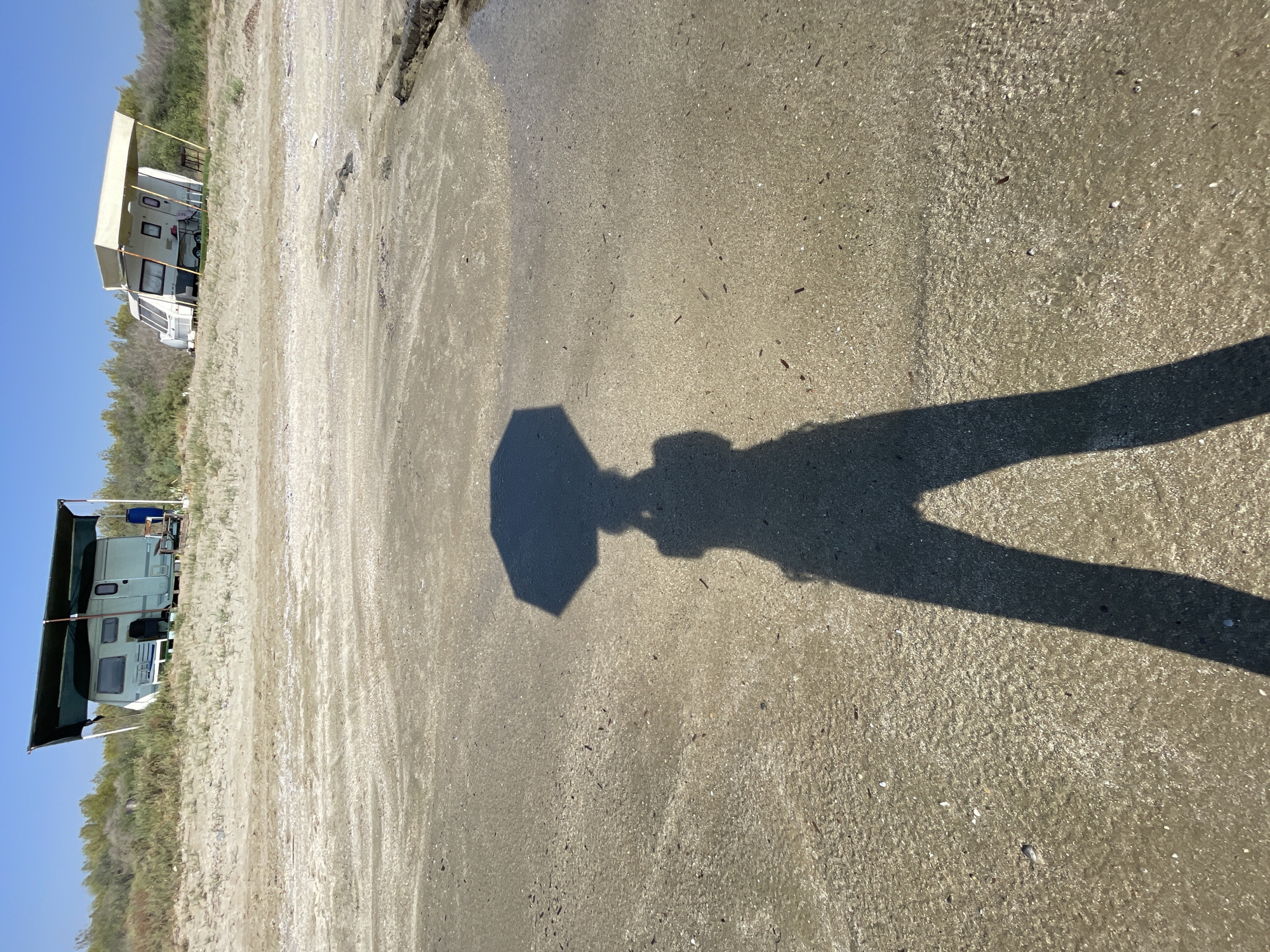 The image depicts a sandy beach area with two small recreational vehicles, or campers, situated on a sandy embankment in the background. The campers are positioned beneath simple canopy structures. The foreground shows the sandy ground of the beach, wet in some areas, with scattered debris. A prominent shadow of a person holding an umbrella is cast on the sand, extending from the lower center of the frame upwards, obscuring the person's face and body but clearly showing their legs and the shape of the umbrella. The sky is clear and blue. The overall scene suggests a sunny day at a coastal location, possibly a campsite, with a person present and casting a shadow on the beach. The location is specified as Agios Sergios, Northern Cyprus / Cyprus.