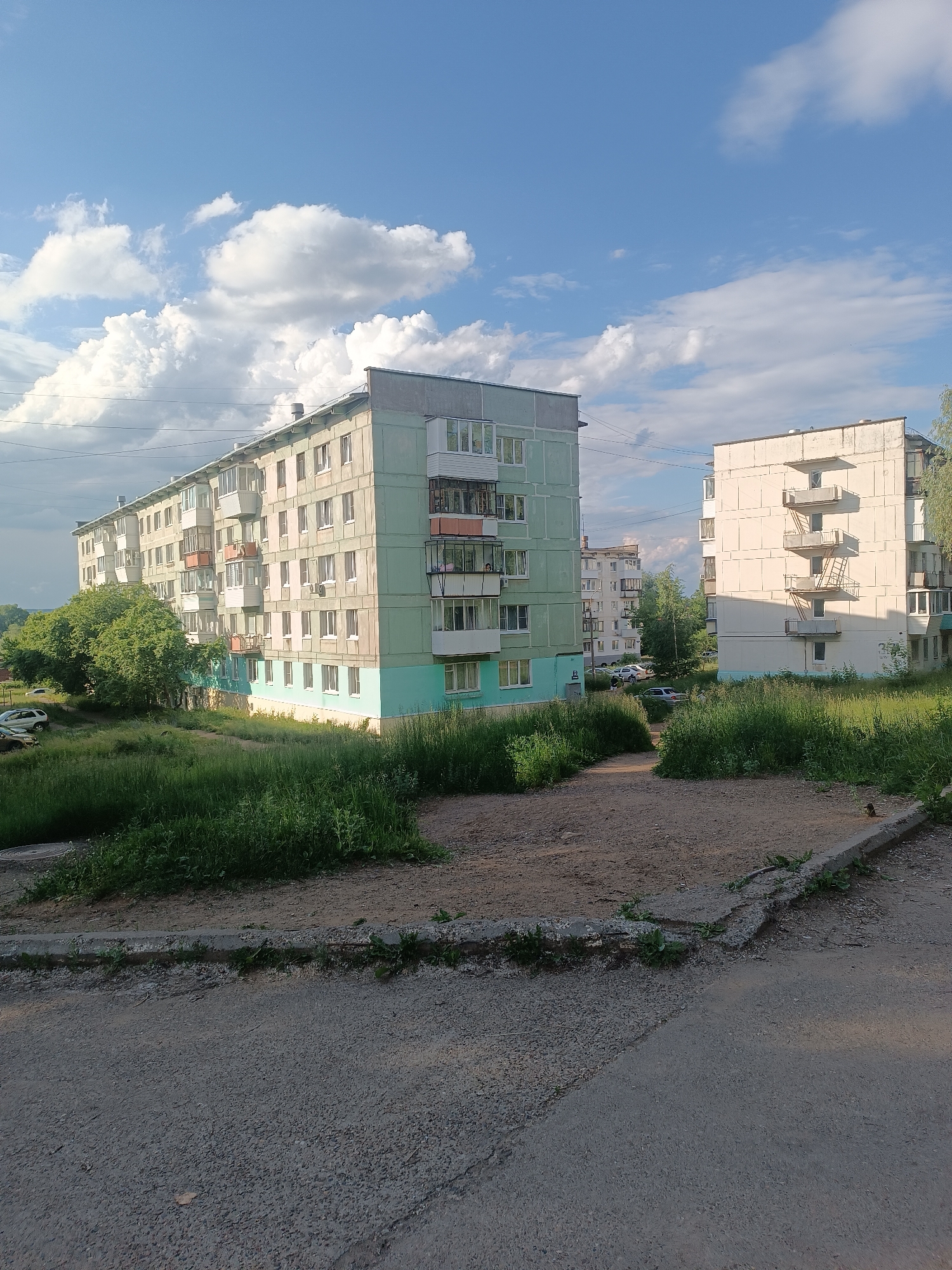 This media file depicts an outdoor scene in Chaikovsky, Russia, on a clear day with scattered white clouds. The time of day appears to be late afternoon or early evening, as indicated by the warm, soft lighting. The setting is a residential area with multiple apartment buildings.
The main subjects are two multi-story apartment buildings. The building on the left is a long, light green structure with multiple floors, balconies, and windows. Trees and dense green grass surround the base of this building. The building on the right is a lighter cream or beige color, also multi-story, with visible balconies and windows, and what appear to be external fire escapes. This building is partially shaded by trees on its right side. In the background, another apartment building with a similar architectural style is visible. The foreground shows a paved area, possibly a road or walkway, with patches of dirt and grass. Several cars are parked in the middle ground, near the apartment buildings.
The scene appears calm and ordinary, suggesting a typical day in this residential neighborhood. There is no discernible activity or event taking place; it is a static view of the environment. Notable details include the well-maintained but somewhat dated appearance of the buildings, the lush green vegetation, and the clear blue sky with fluffy clouds, indicating pleasant weather. No visible text is present in the image.