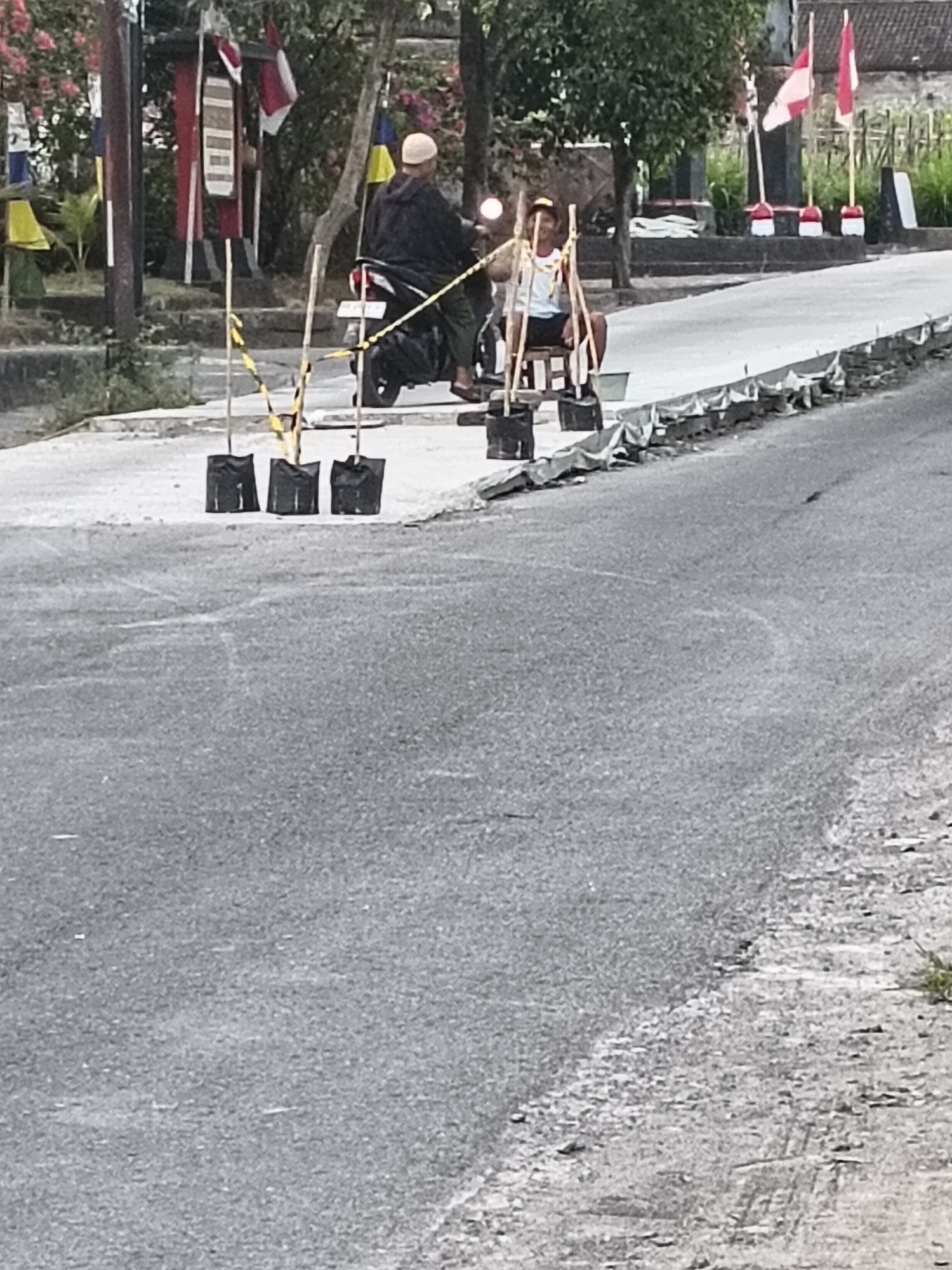 The image depicts a street scene in Sukorini, Indonesia, with a focus on a motorcycle and a makeshift cart. A man wearing a dark jacket and a white cap is riding a black motorcycle. He is followed by a wooden cart, also made of wood, carrying a young boy wearing a white sleeveless shirt and a dark cap. The cart appears to be attached to the back of the motorcycle. The road is paved and gray, with some debris and tire marks visible.
Along the side of the road, several black plastic bags are placed upright, supported by bamboo stakes. Yellow and black caution tape is strung between some of these stakes. In the background, there are trees, a structure with flags, and a building with a tiled roof. The flags are red and white, consistent with the Indonesian flag. The overall impression is of a somewhat cluttered, possibly rural or semi-urban environment. The lighting suggests it is daytime.