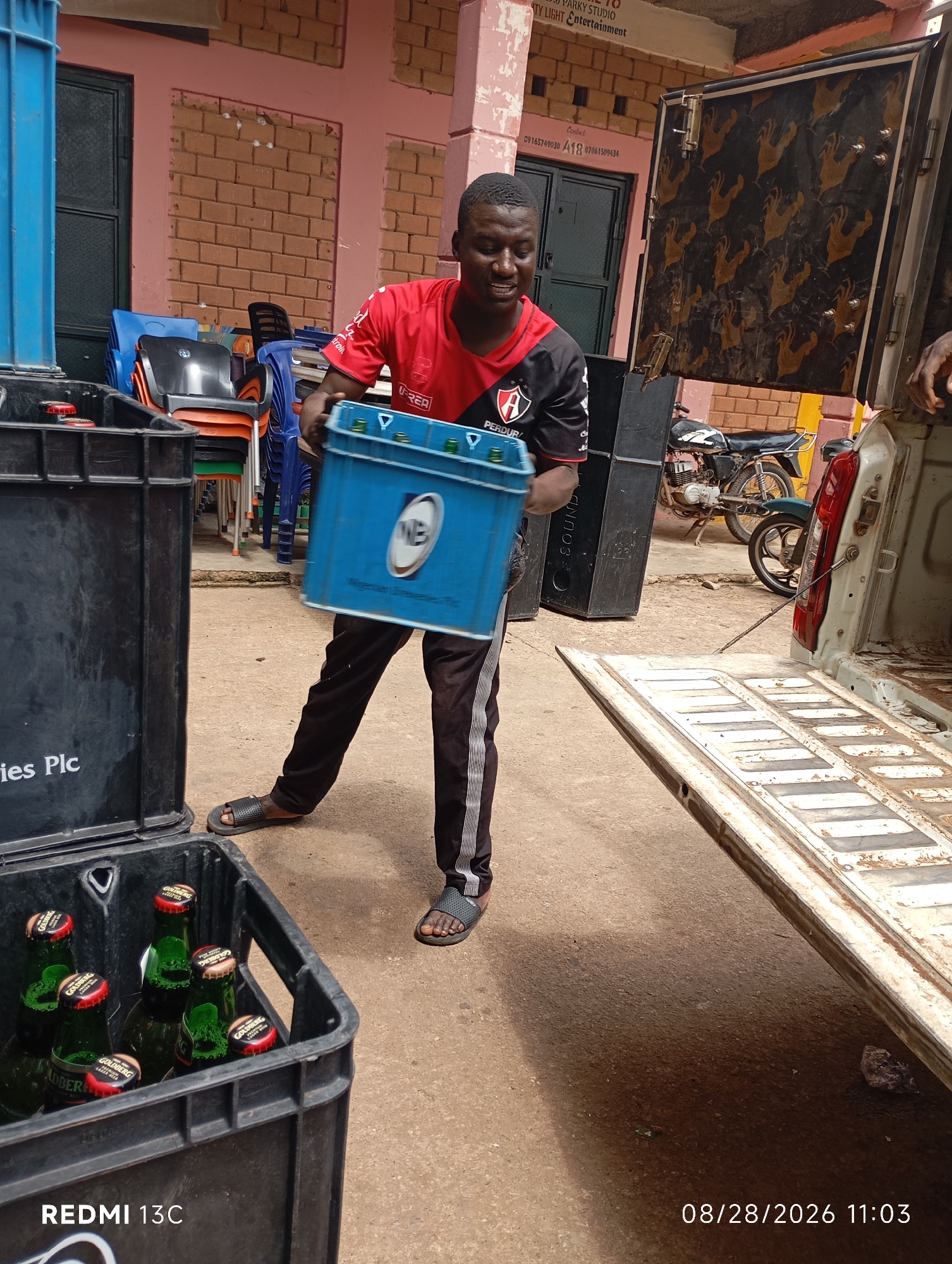 In Shendam, Nigeria, a young man in a red and black jersey with the logo "A PERDUR..." on it is carrying a blue crate containing bottles. He is wearing black pants with white stripes and black sandals. The scene appears to be an outdoor loading area, possibly for a distribution business. A white truck with its tailgate down is visible to the right, with a motorcycle parked behind it. Black crates filled with green beer bottles are in the foreground. The environment features a brick building with pink trim, and stacked chairs. A black object with a rooster pattern is attached to the truck. The date and time stamp indicate August 28, 2026, at 11:03 AM.