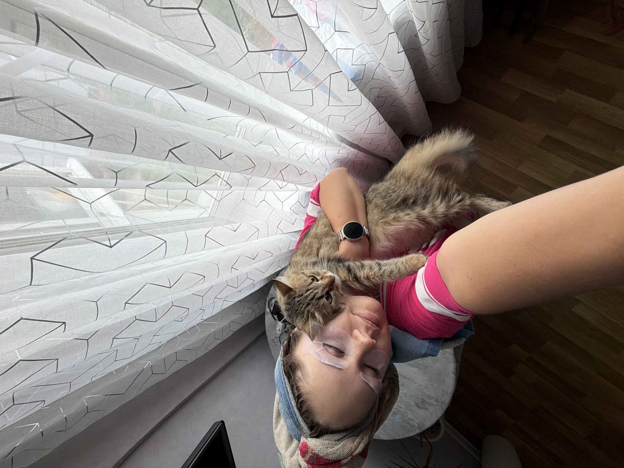 A young woman with a towel wrapped around her head and under-eye patches is lying down, holding a cat. She is wearing a pink and white striped shirt. The cat is brown and grey tabby and is resting on her chest. The background shows a window with a patterned white curtain. The scene appears to be indoors, possibly in a home. There is no specific indication of the location being in Chaikovsky, Russia.