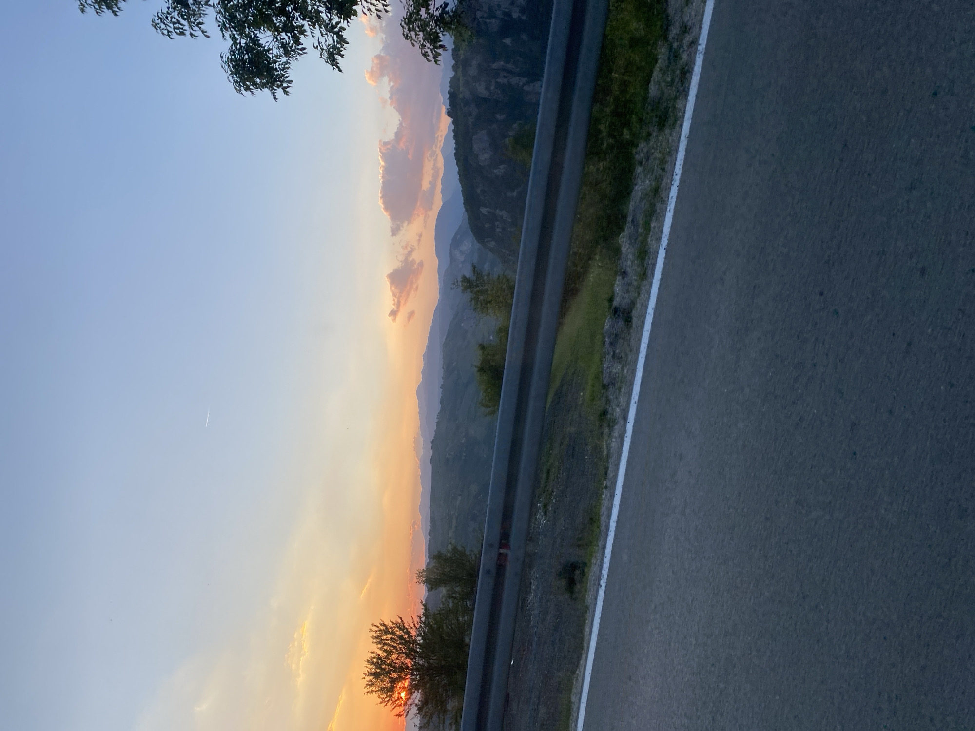 The image captures a scenic mountain landscape during sunset, viewed from a roadside. In the foreground, a paved road with a white line marking its edge stretches into the frame. A metal guardrail runs along the side of the road, separating it from a sloped embankment covered in grass and some scattered rocks. Beyond the guardrail, the terrain descends into a valley characterized by rolling hills and dense vegetation. Further in the distance, layered mountain ranges fade into the horizon, under a sky painted with warm hues of orange, yellow, and pink, indicative of the setting sun. A few wispy clouds are present, and the sun itself is partially visible, peeking through a tree on the left side of the image. The overall scene suggests a natural, undeveloped environment, likely in a rural mountainous region like Šavnik, Montenegro. There are no people or visible signs of urban development. The time of day is clearly sunset.