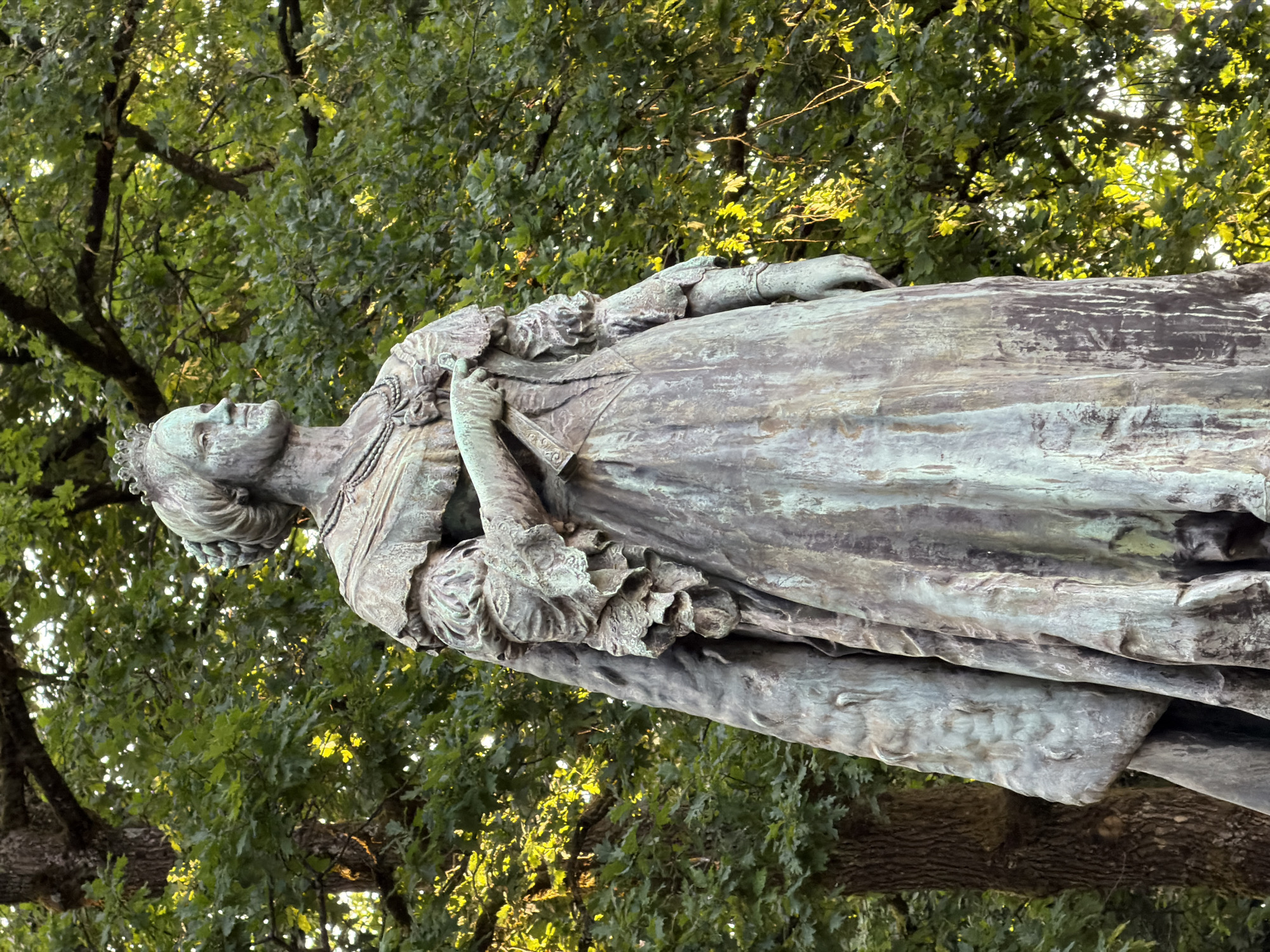 A full-body bronze statue of a woman in regal attire stands outdoors against a backdrop of lush green trees. The statue depicts the woman wearing a crown, a detailed necklace, and a long, flowing gown with ornate sleeves and a cape. Her left hand is positioned near her chest, holding a rolled document or fabric. The bronze has a weathered patina, with visible green and dark tones. The background consists of a dense canopy of oak trees, with sunlight filtering through the leaves, creating a dappled effect and highlighting parts of the statue and foliage. The statue appears to be situated in a park or garden area. The statue is located in the Parc de la Pétrusse in Luxembourg City, Luxembourg.