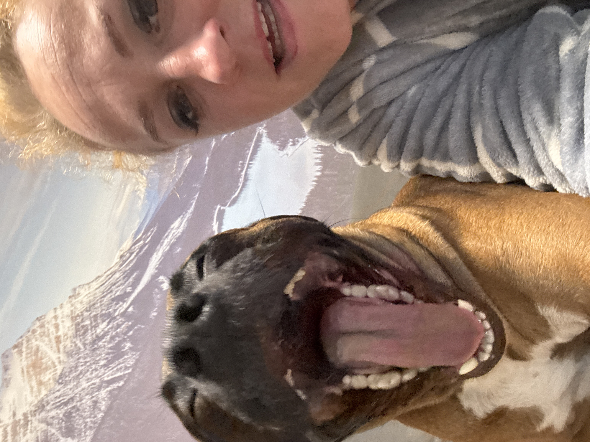 The image shows a woman and a dog in a selfie, taken at a close, upward angle. The woman is at the top of the frame, her face partially visible, and she appears to be smiling. She has blonde hair and is wearing a grey and white striped top. The dog is positioned below her, and the photo mainly captures the inside of its mouth, including the tongue and teeth. The dog's fur is brown and white. The background shows a blurry image that resembles mountains. There is no information in the image to suggest it was taken in the city of Chaikovsky, Russia.