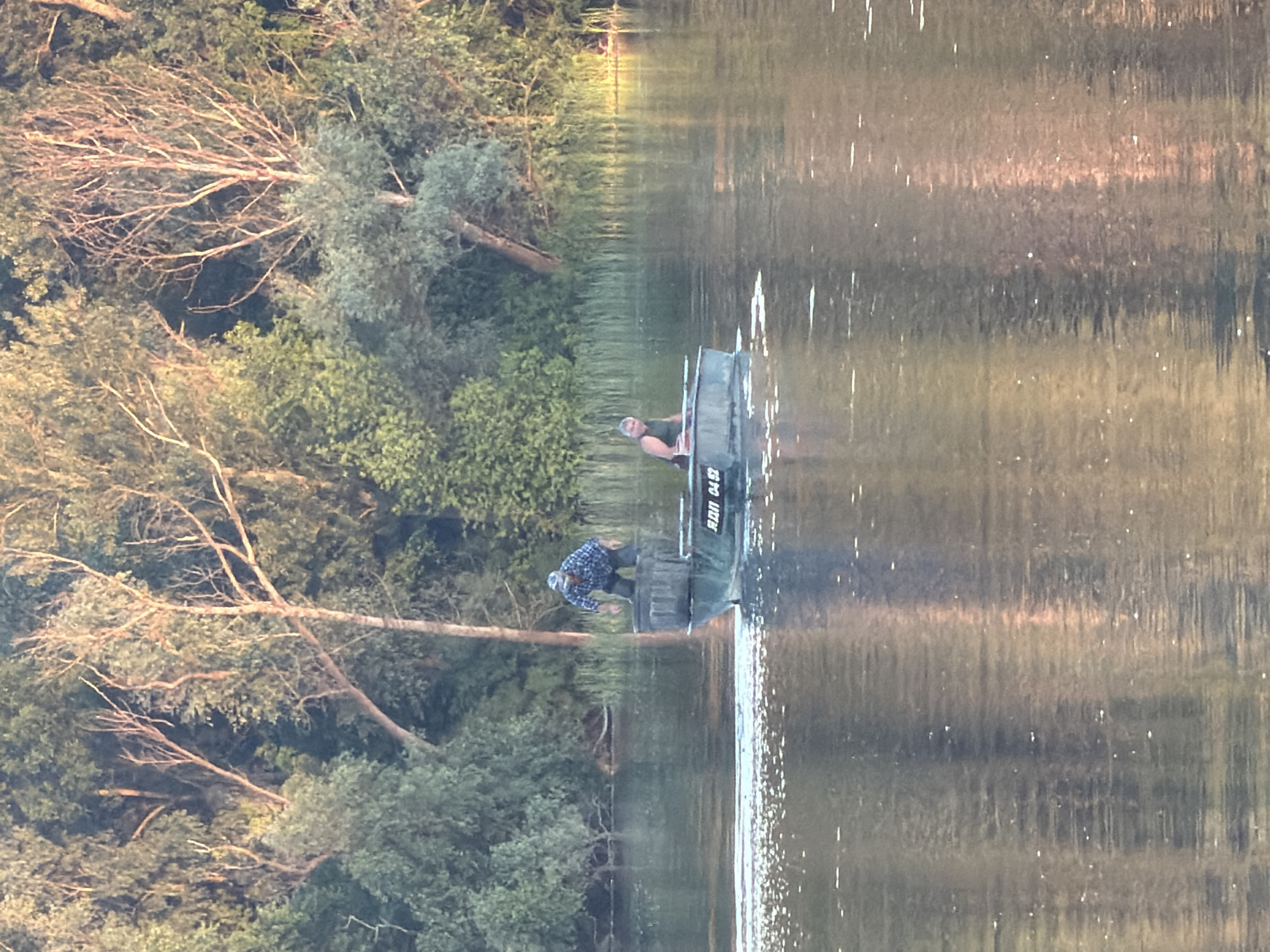 In this image, two individuals are in a small boat on a calm body of water, likely a river or lake, near a wooded shoreline. The scene is oriented sideways. The boat is dark-colored and appears to be a jon boat. One person, wearing a plaid shirt, is seated in the boat, facing forward and looking towards the right side of the frame. Another person, wearing a dark shirt, is standing in the boat, leaning over the side and appears to be reaching into the water.
The setting is lush and green, with dense trees and foliage lining the bank. Several branches from trees overhang the water, casting dappled shadows. The water's surface reflects the surrounding trees and the sky, creating a blurred, impressionistic effect. Small white highlights on the water suggest either ripples from the boat or reflections of sunlight. The overall lighting suggests it is either early morning or late afternoon, with a warm, soft light illuminating the scene.
On the side of the boat, some text is visible, appearing to read "RDN 04 52". The presence of trees and the type of boat suggest a rural or natural environment, possibly a recreational area. The individuals' attire does not indicate any specific activity beyond being on the water. The mood of the image is peaceful and serene, emphasizing the natural beauty of the location. The location context of Obukhivka, Ukraine, places this scene in Eastern Europe.