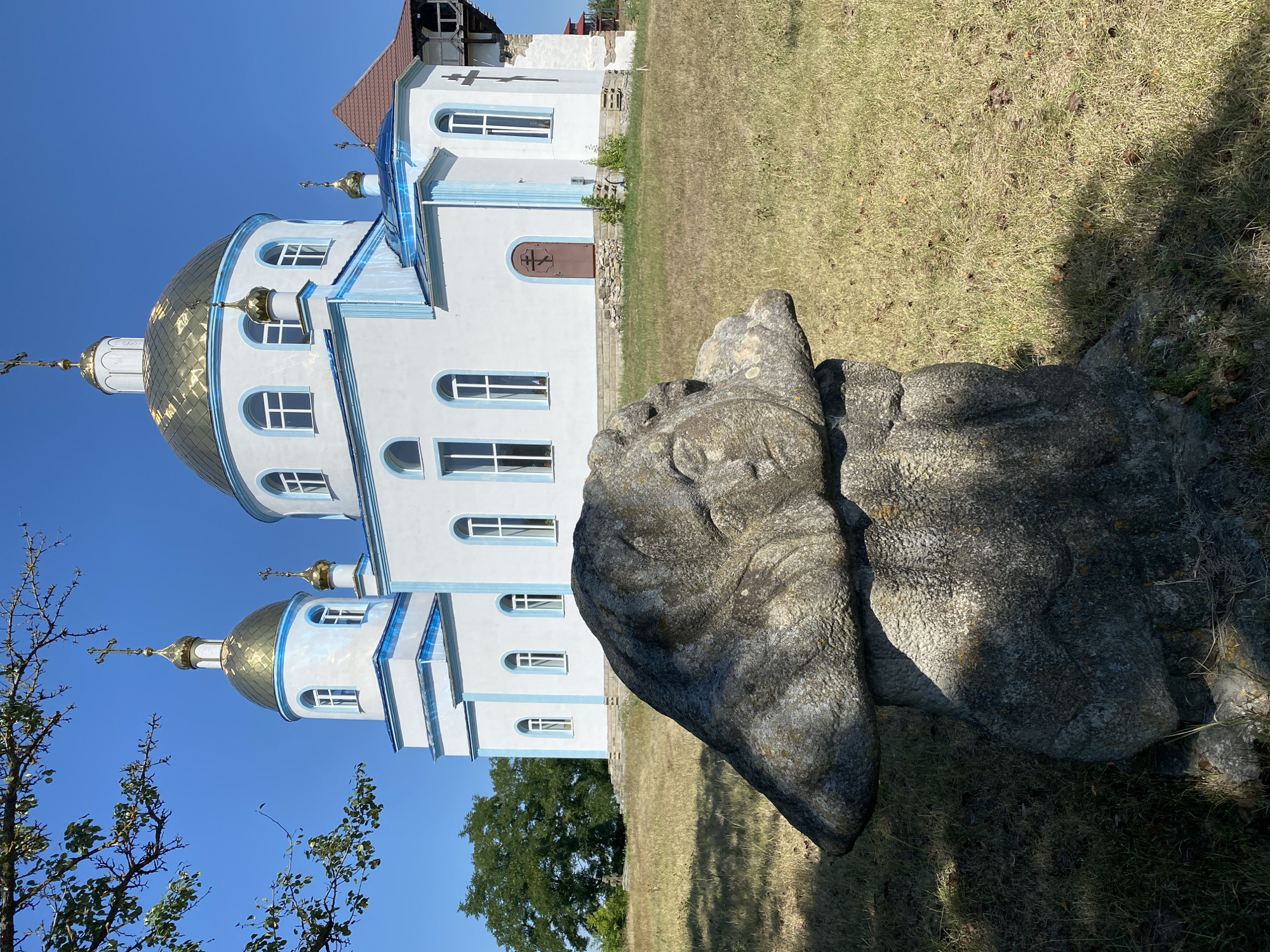 The image shows a stone sculpture in the foreground and a white church with golden domes in the background. The sculpture appears to be weathered and is positioned on a grassy area. The church is a prominent structure with arched windows and blue trim. The sky is clear and blue. The scene is located in the city of Busha, Ukraine.