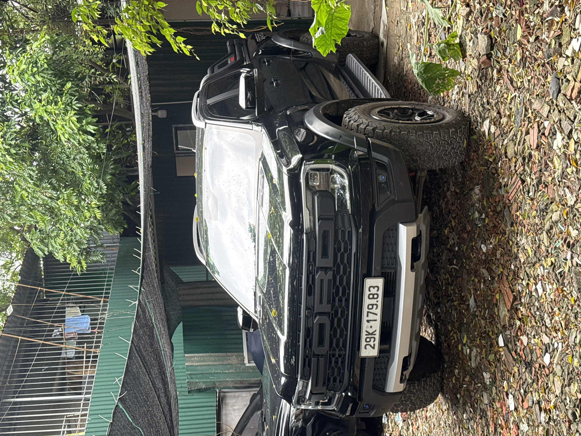 The image shows a black Ford Raptor pickup truck parked on a surface covered with fallen leaves, possibly near a building. The truck's front license plate is visible and reads "29K-179.83." The surrounding environment includes foliage, indicating a somewhat natural setting. The presence of a building and a fence suggest an urban or suburban location. Based on the license plate prefix "29K," it's likely the location is in Hà Nội, Vietnam. There is no presence of people, and the focus is solely on the vehicle and its immediate surroundings.
