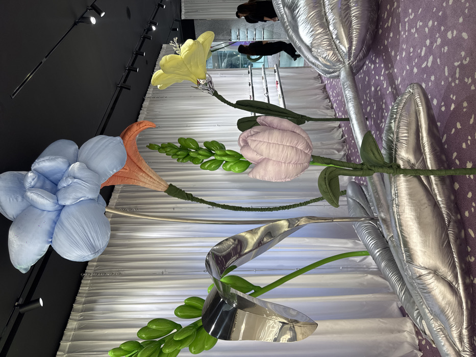 The image depicts the interior of a Moncler Genius retail store located in Siam Paragon, Bangkok, Thailand. The scene is dominated by an immersive art installation featuring oversized, plush, and metallic floral sculptures.
In the foreground and midground, several large, fabric-wrapped flowers are prominent: a light blue multi-petaled flower, a vibrant yellow flower with silver metallic accents, and a light pink tulip-shaped flower. These are accompanied by large, glossy green plant stalks and leaves, as well as expansive silver metallic, inflatable plant elements resembling large leaves and bulbs resting on the patterned purple carpet. The stems of the plush flowers are wrapped in dark green fabric.
The floor is covered with a purple carpet featuring irregular white speckles. The ceiling is black, fitted with numerous black track lights. The background consists of white, pleated curtains that run along a wall, behind which glass display shelves stocked with merchandise are visible. Two individuals in dark clothing are partially visible in the right background, appearing to observe the displayed items within the retail space.